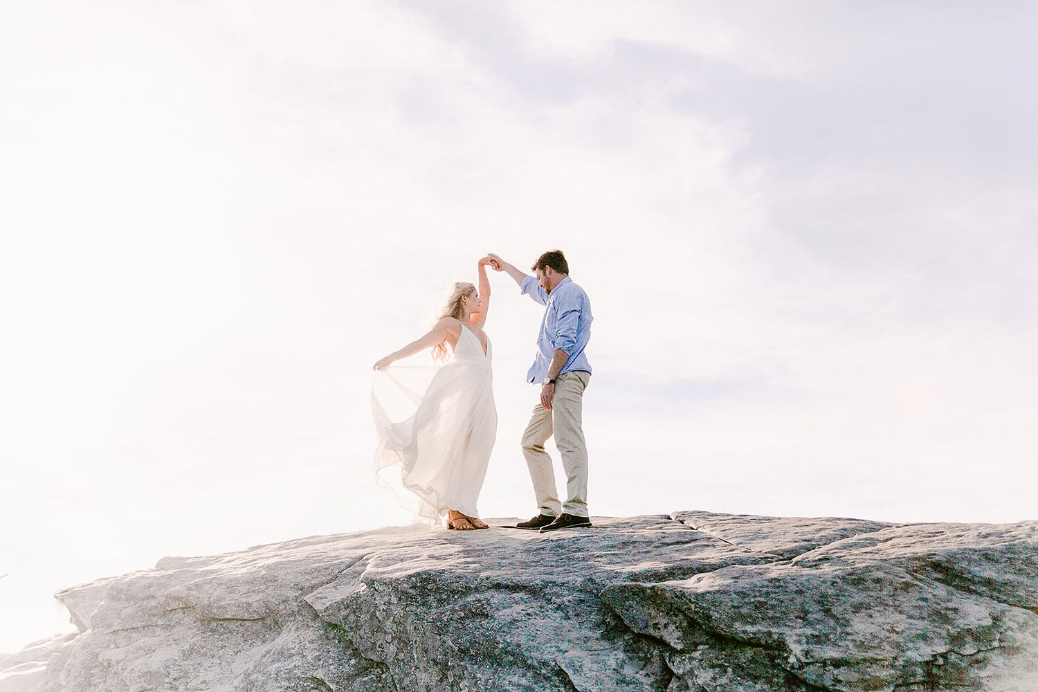 Couple playfully explores mountain terrain while dressed in light summer clothing against cloudy sky.
