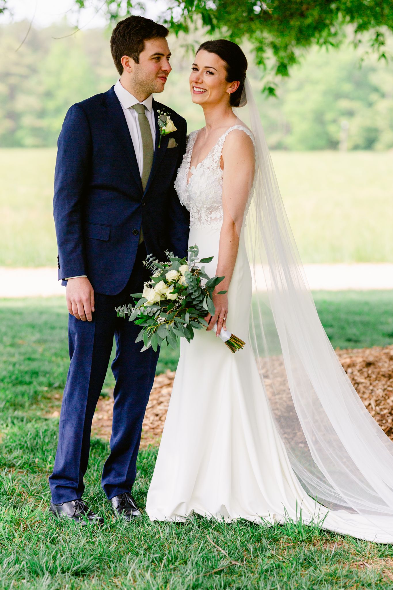 A bride in a lace wedding gown with flowing veil stands with groom in navy suit outdoors under green trees.