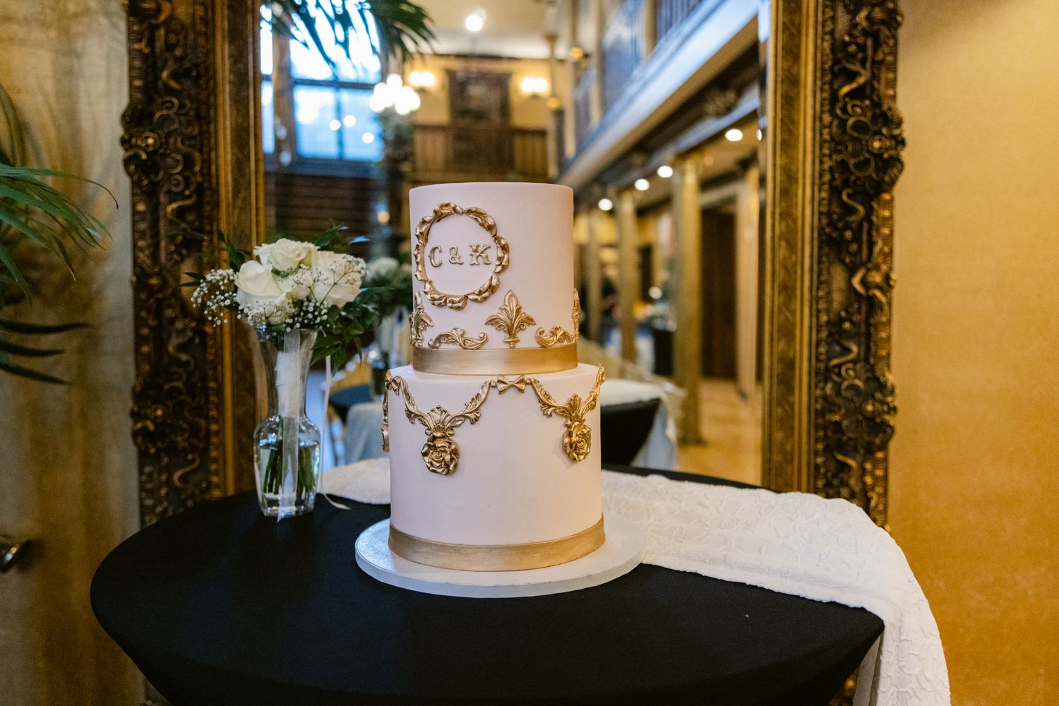 Elegant white wedding cake with delicate gold details and floral accents displayed in an ornate reception venue.