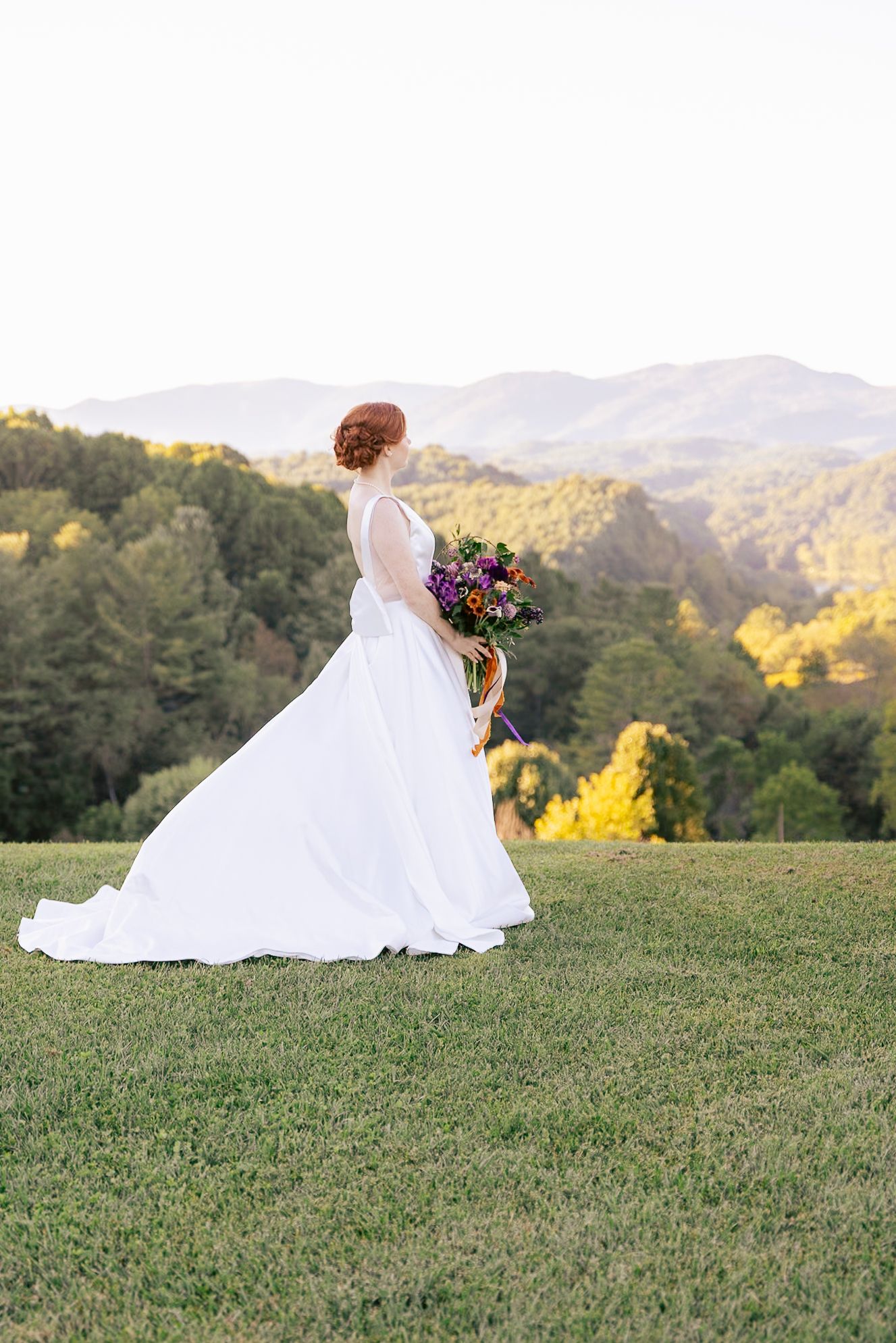 A figure in a flowing white wedding dress sits gracefully on a grassy hilltop overlooking mountains at sunset.