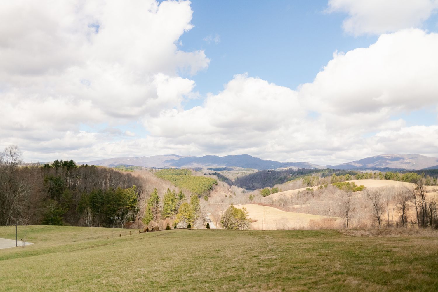 Rolling hills and mountains stretch across a scenic rural landscape under puffy white clouds on a bright day.