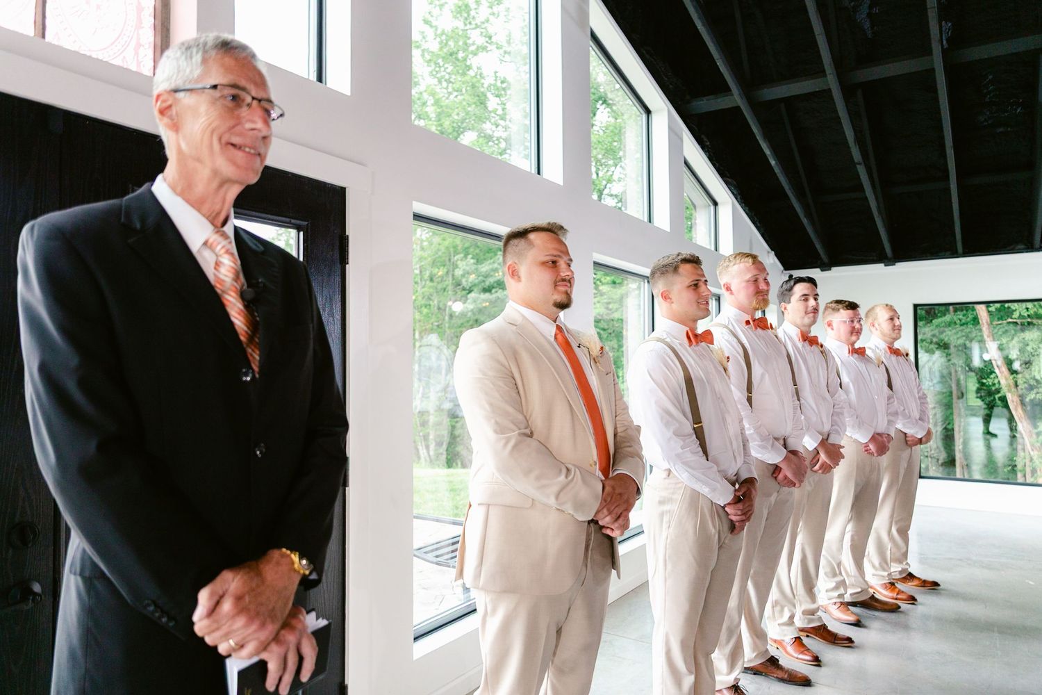 A wedding party lineup of groomsmen in light suits and dark suits stands along a covered outdoor walkway.