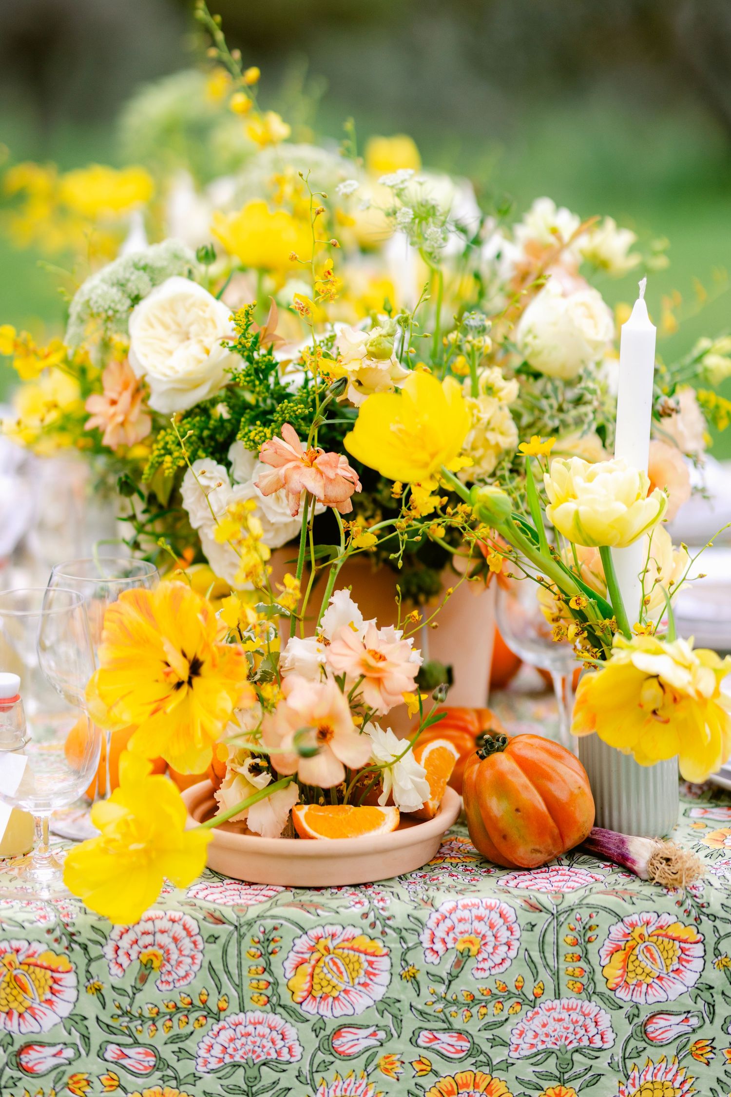 Fall table decor features bright yellow and peach floral centerpieces with small pumpkins on a colorful patterned tablecloth.