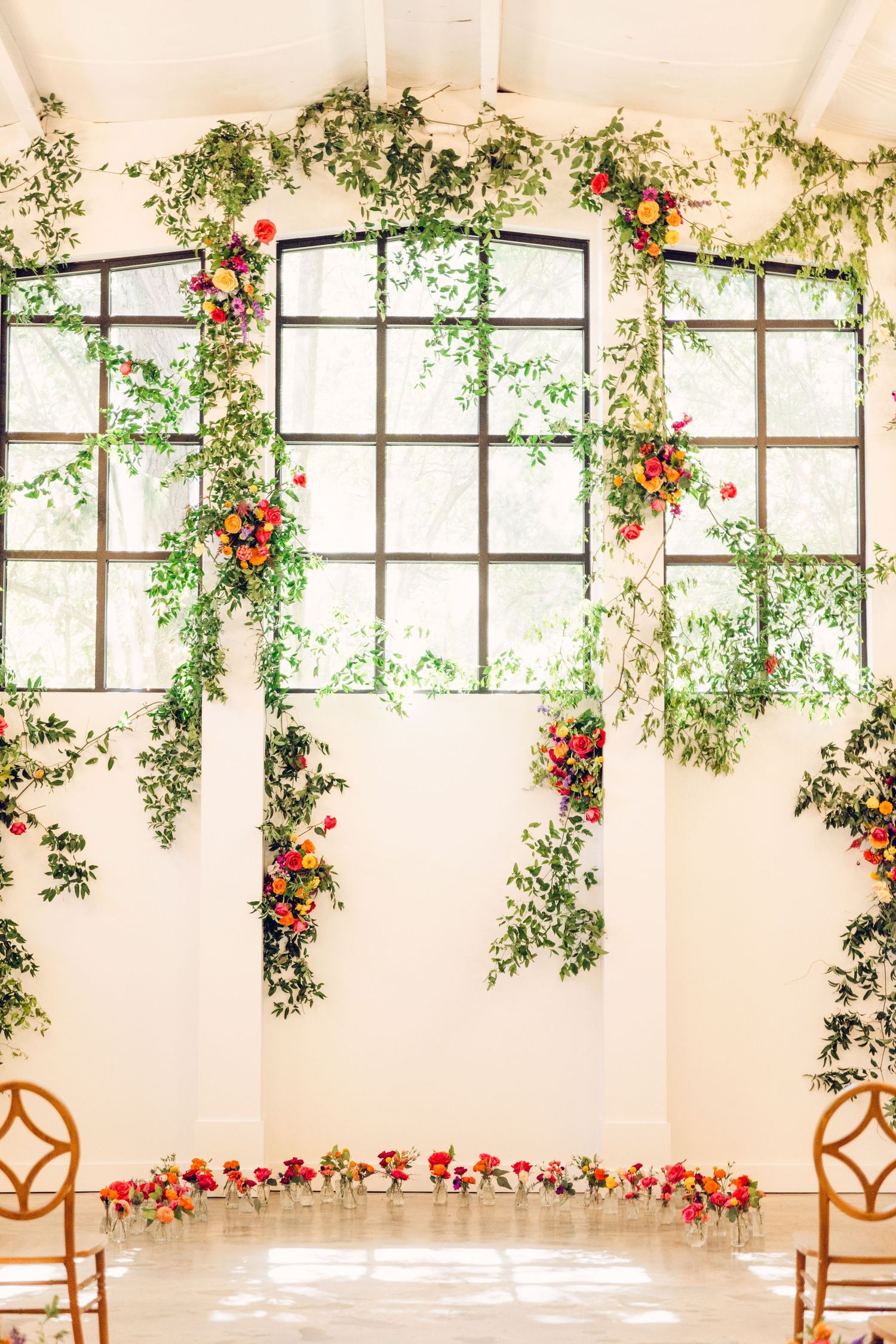 Bright indoor venue with large windows decorated with cascading greenery and red flowers for a romantic wedding ceremony.