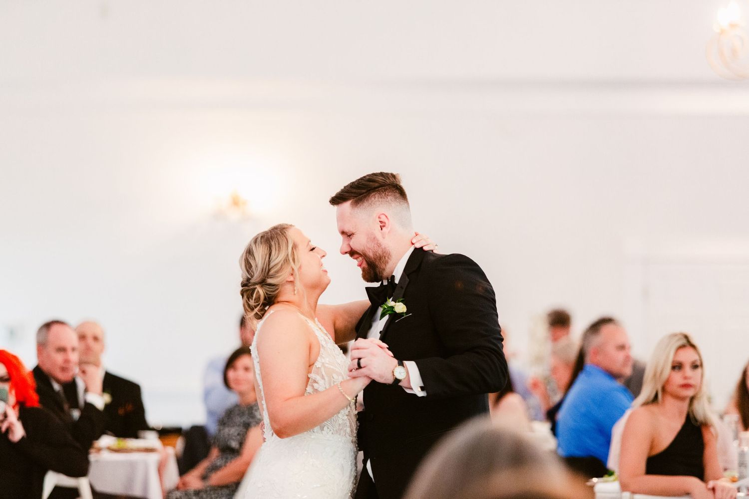 A couple shares a romantic kiss during their wedding reception while guests watch and smile in the background.
