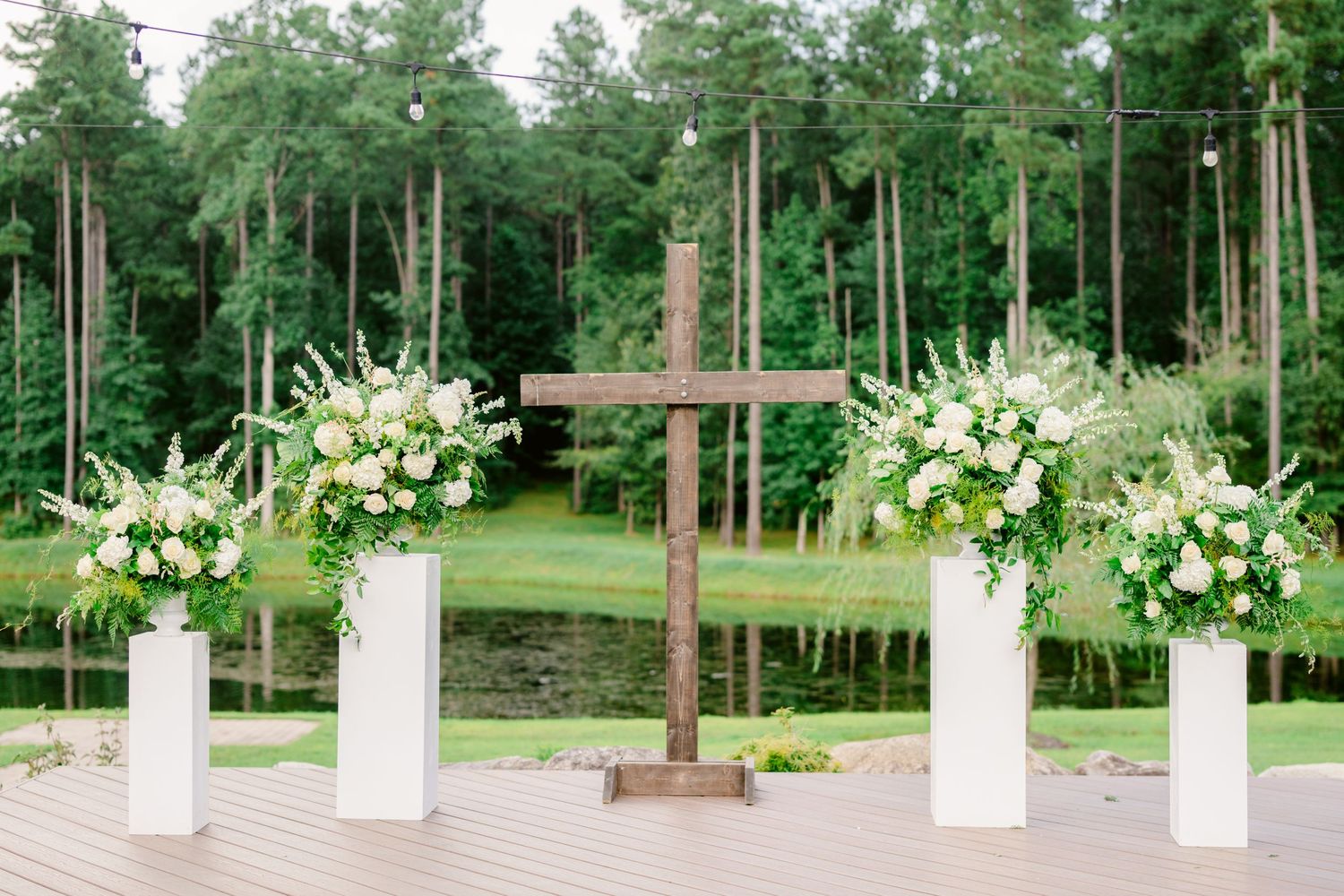 Wooden cross and white floral arrangements on pedestals create an elegant outdoor wedding ceremony setup in a pine forest.