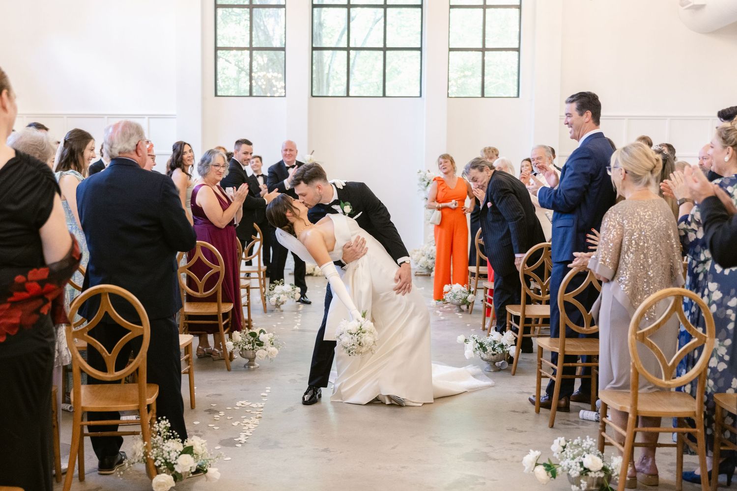 Wedding guests dance and celebrate in a bright white reception venue with rustic wooden chairs and large windows.