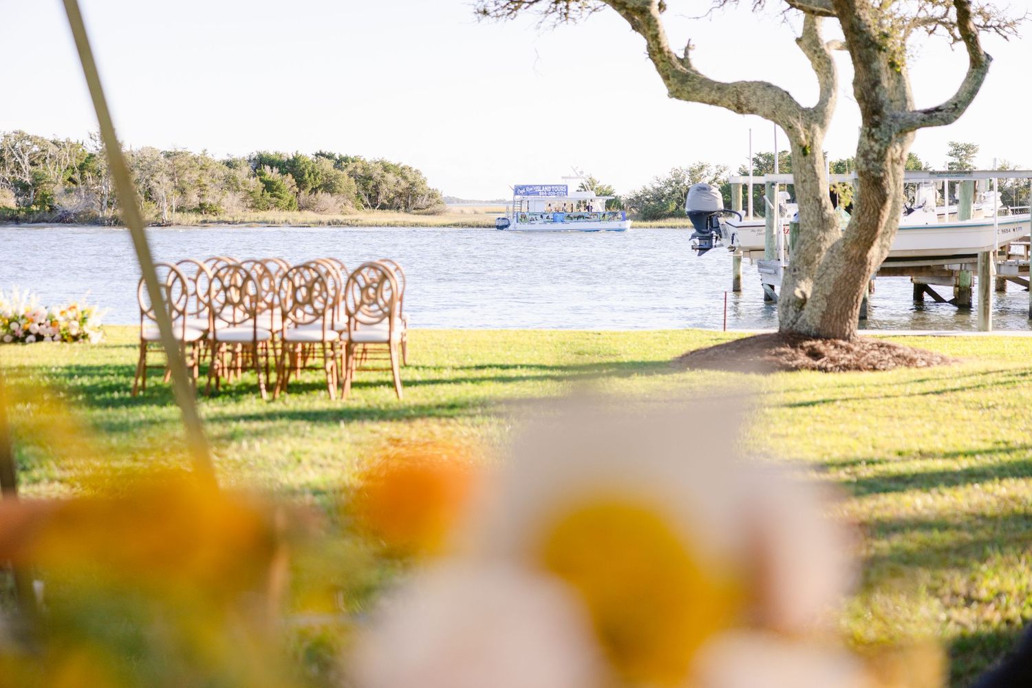 Waterfront wedding venue setup with wooden chairs and water views under twisted trees.