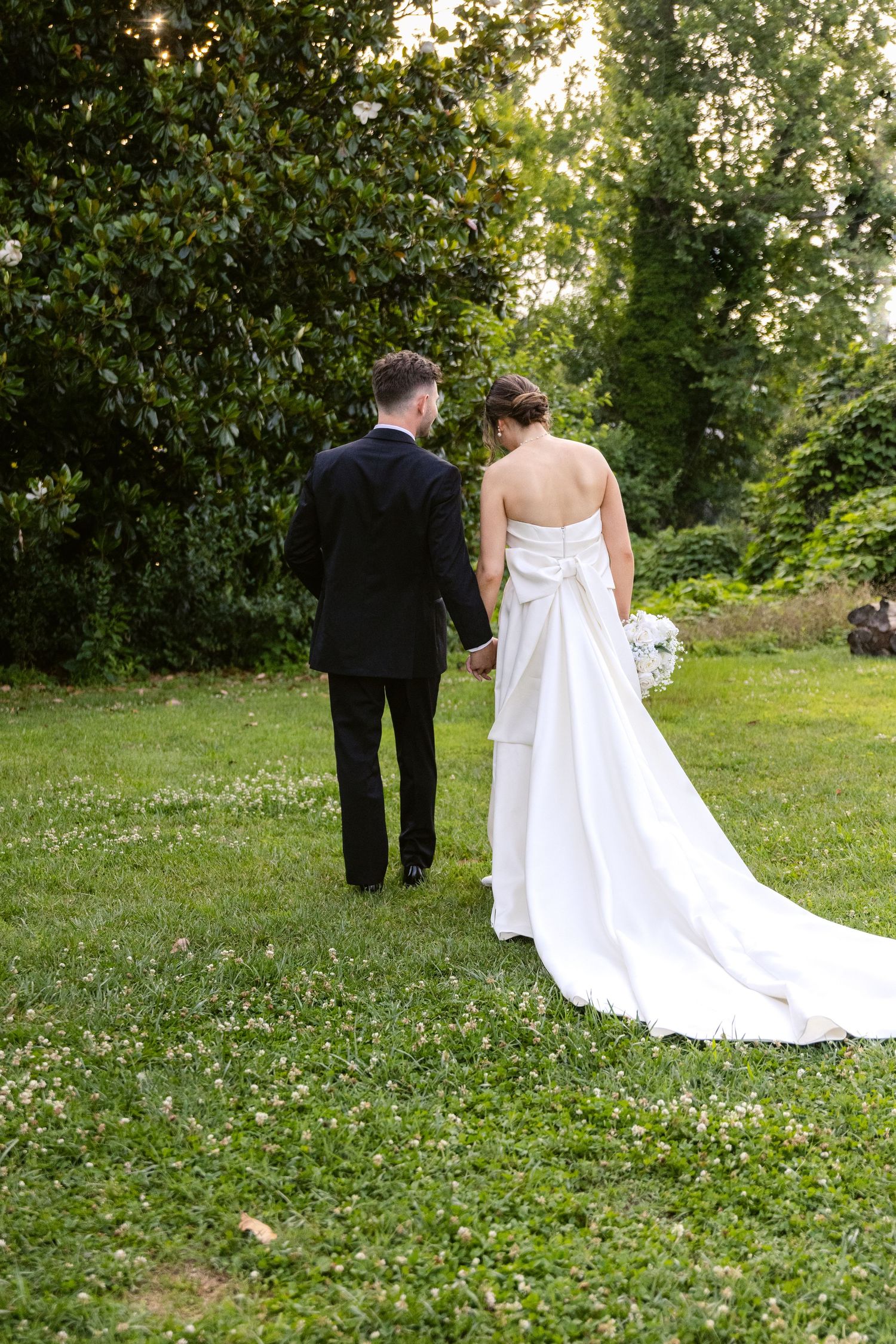 A newlywed couple walks together on a lush green lawn surrounded by trees, the bride wearing a flowing white gown.
