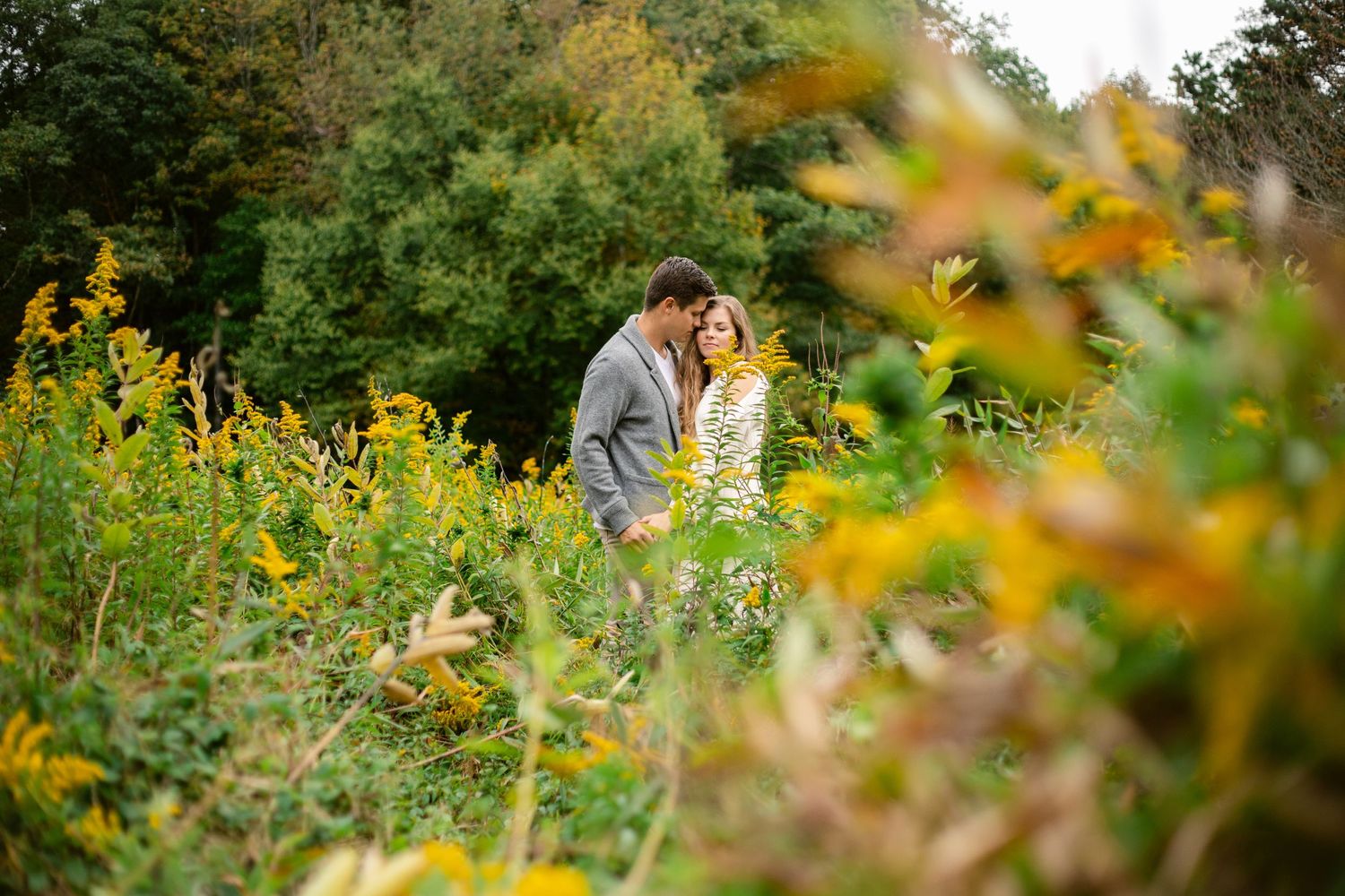 A couple surrounded by yellow wildflowers and autumn foliage during an outdoor photoshoot.