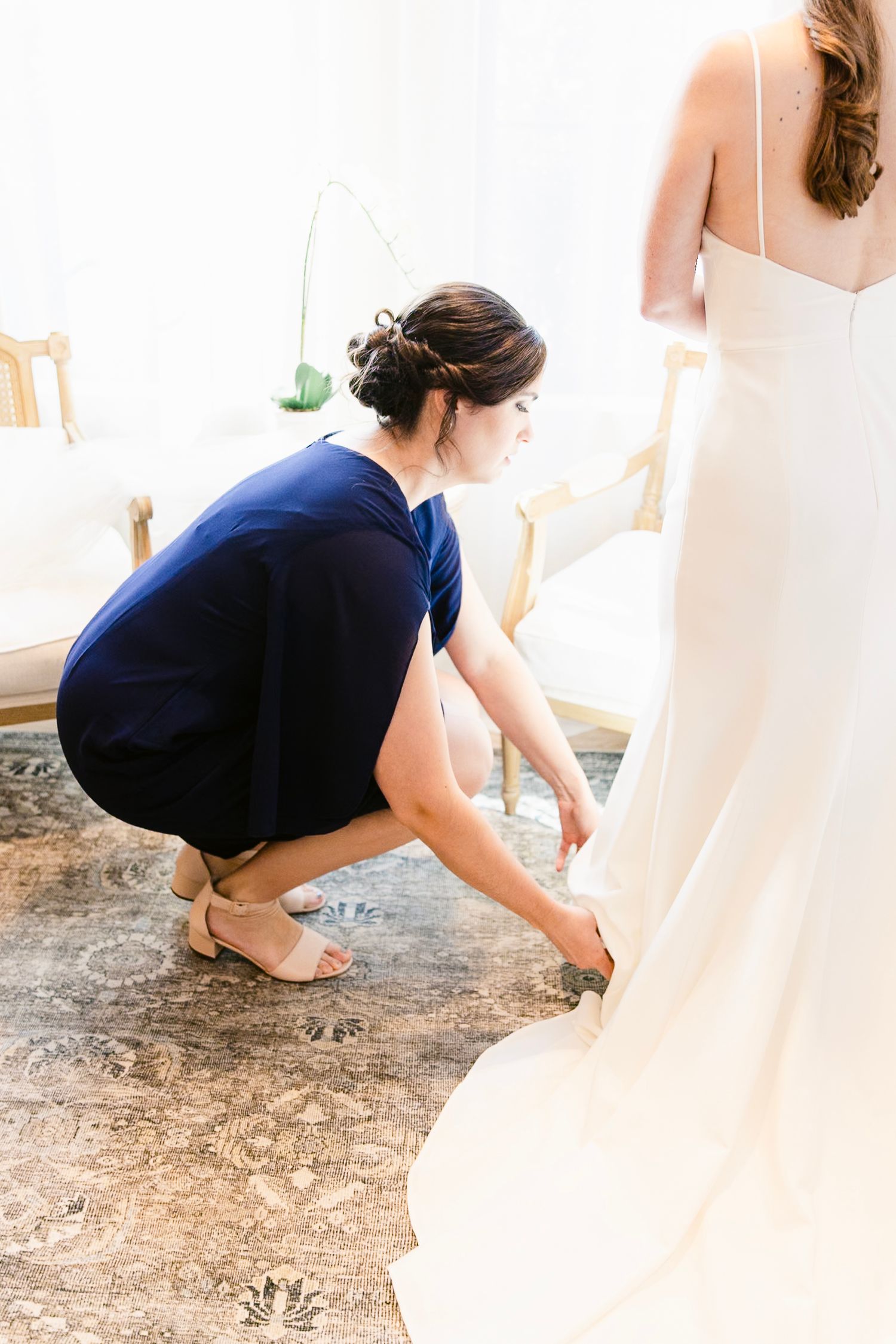 A person in a navy dress adjusts the hem of a flowing white wedding gown on a patterned carpet.