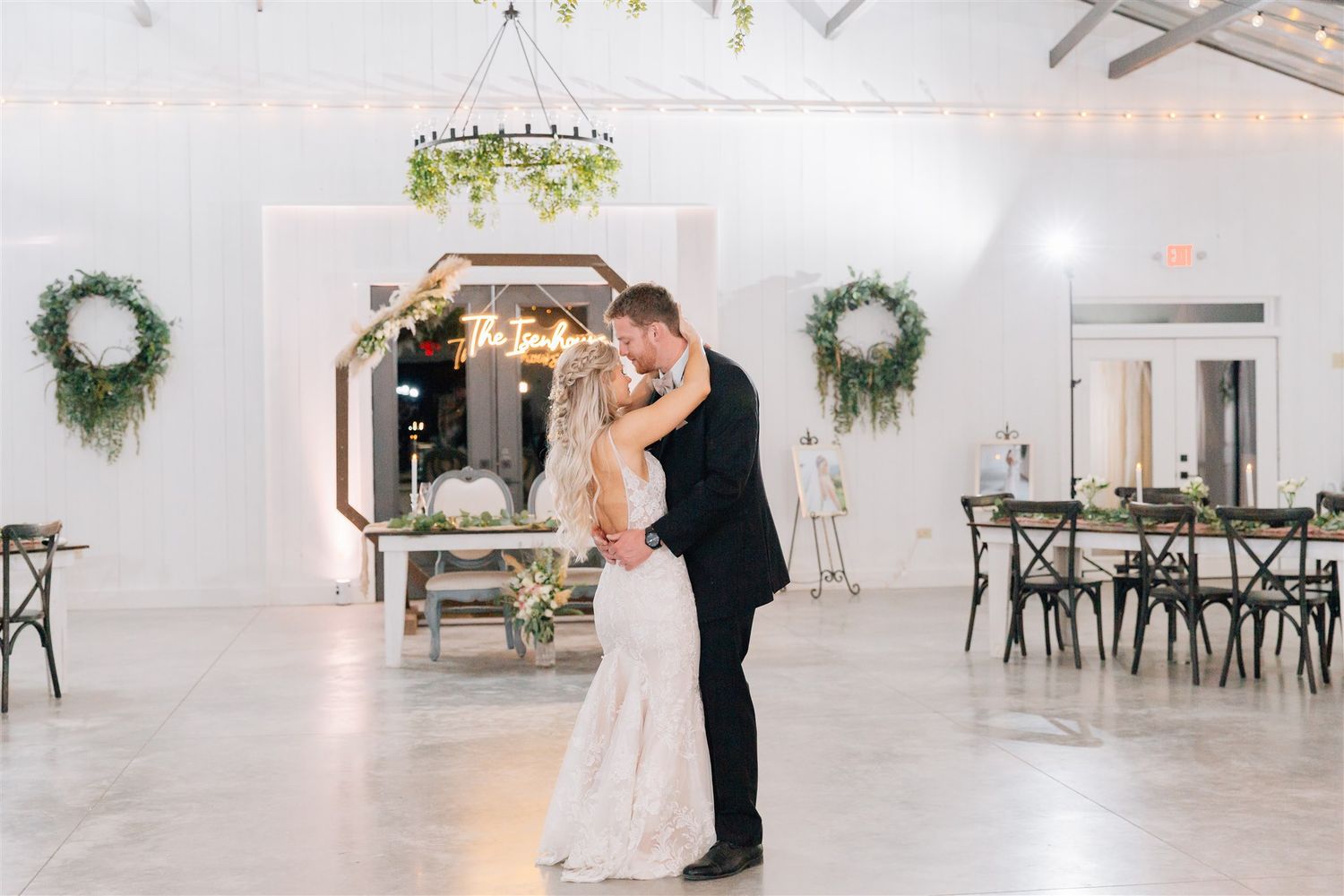 A couple shares their first dance in an elegant white reception venue decorated with greenery and chandeliers.