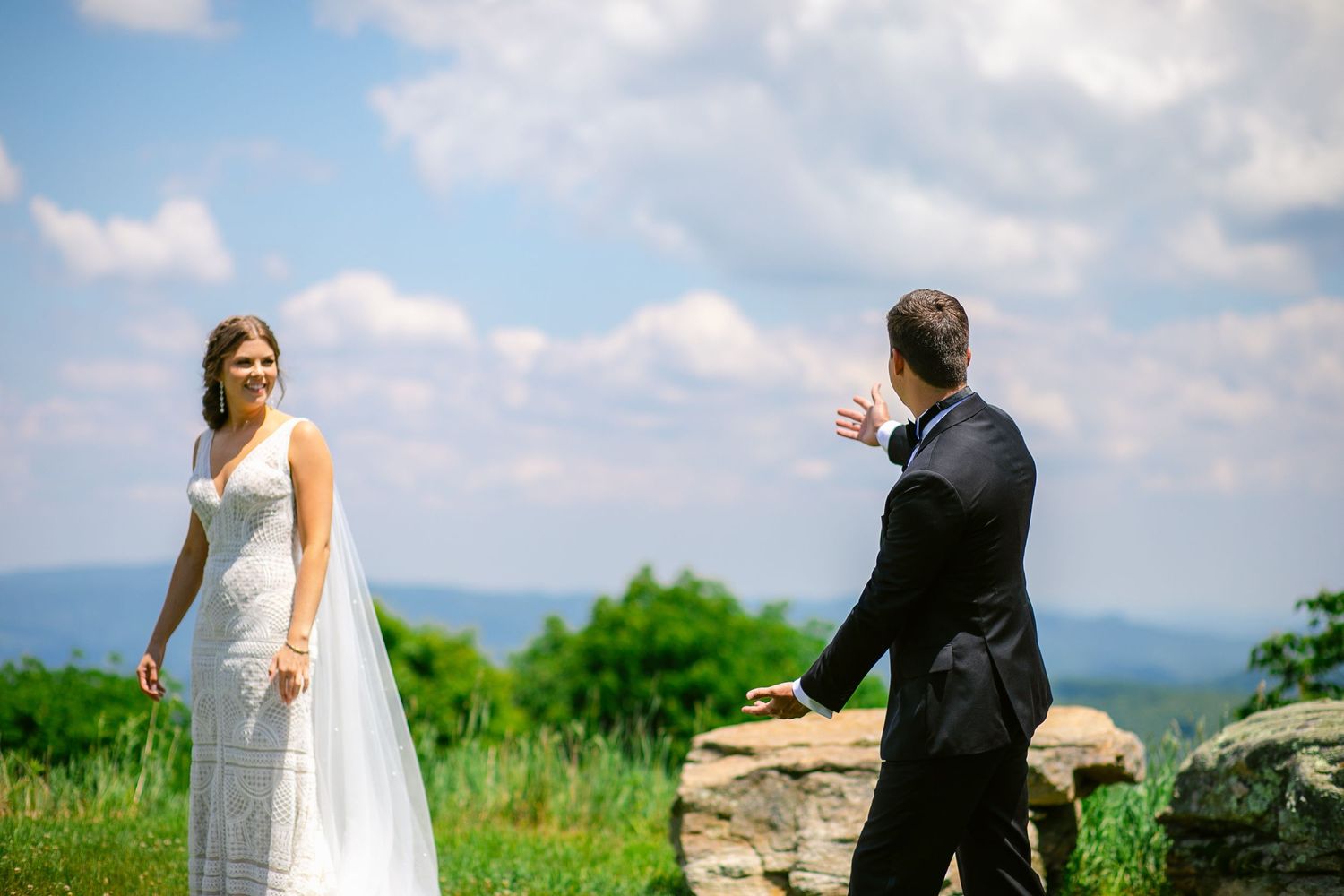 A couple in wedding attire stands on a grassy hilltop overlooking a scenic mountain landscape on their special day.