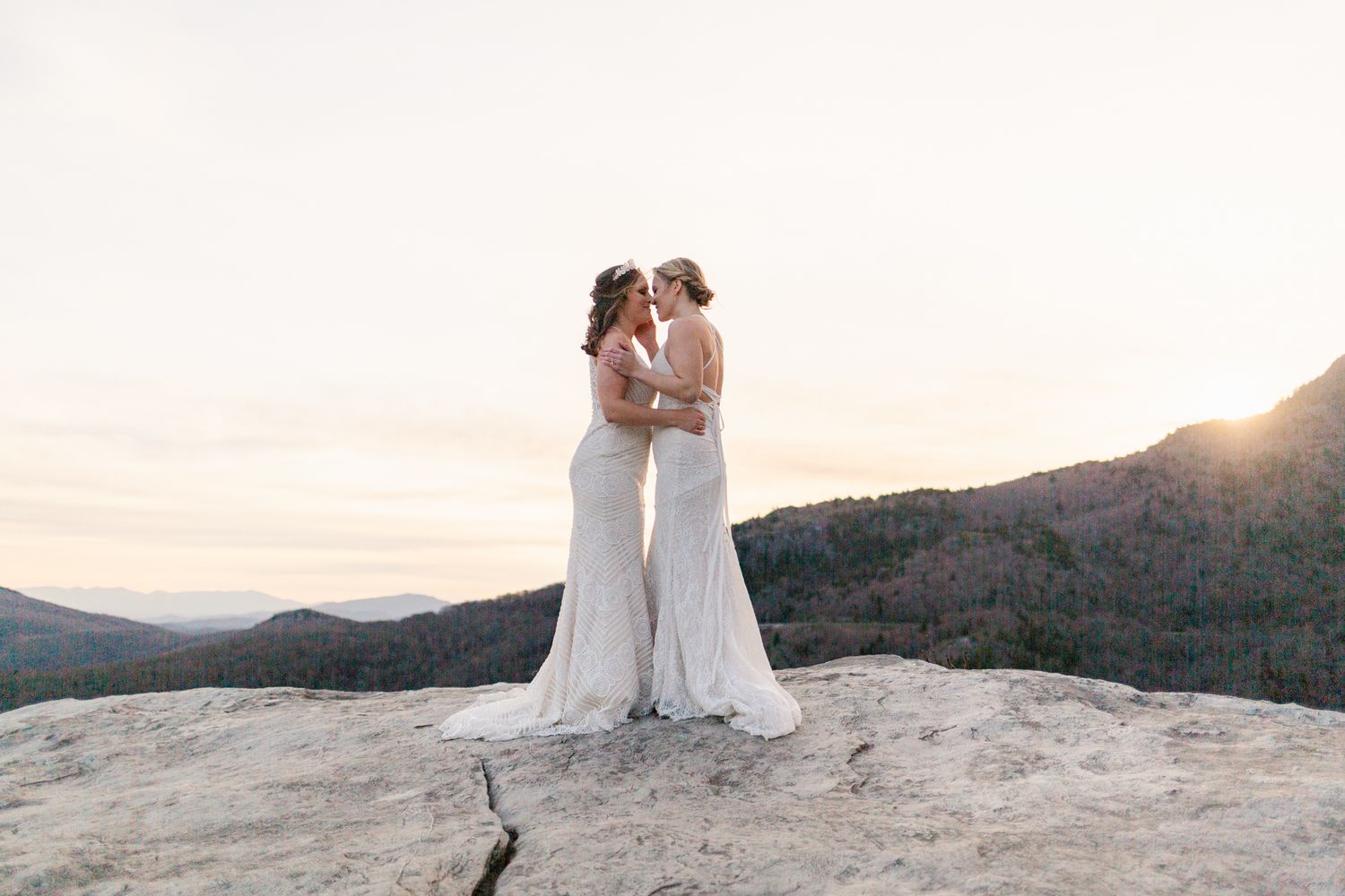 Two people share an intimate moment at sunset on a mountain overlook with dramatic views of distant peaks.