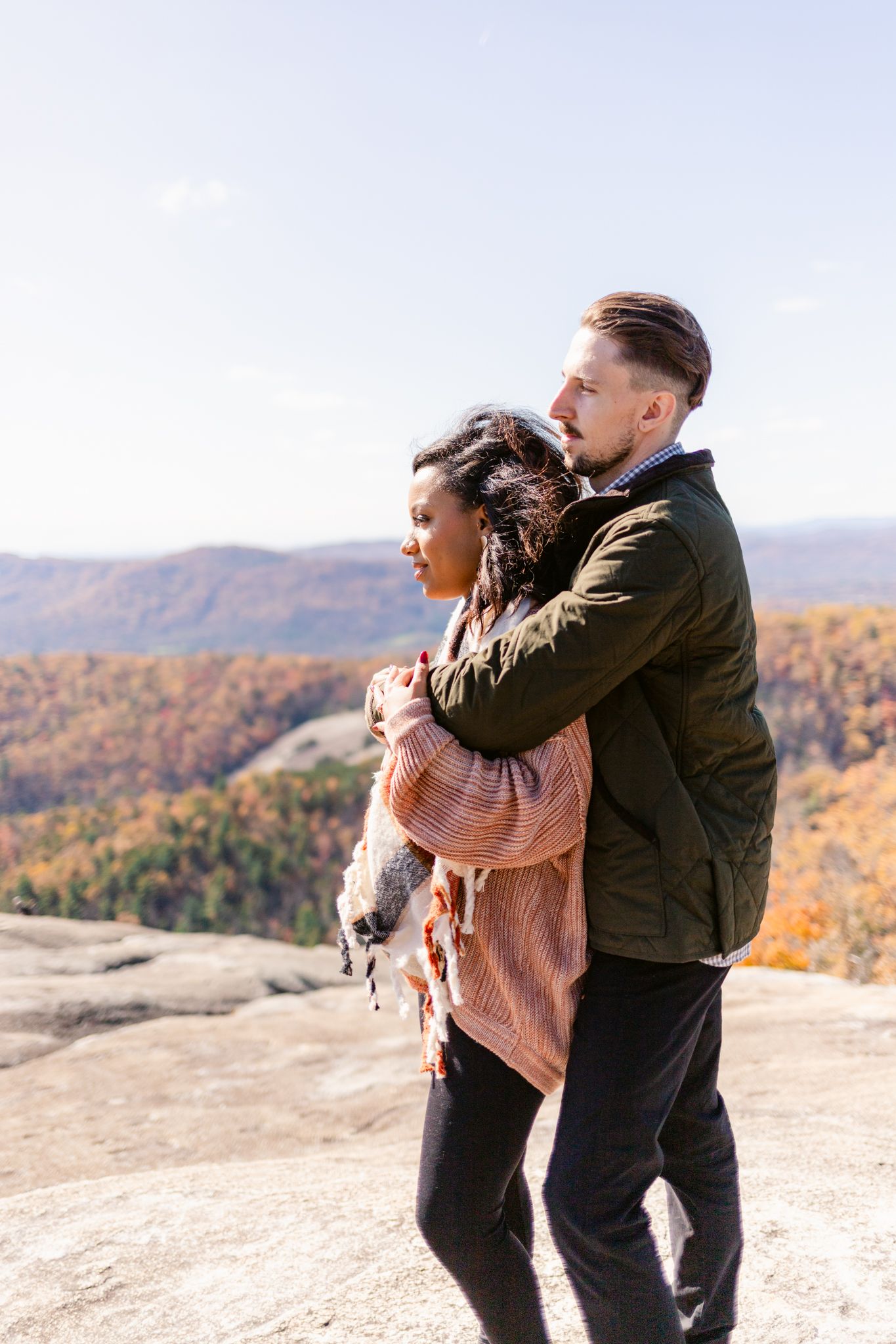 A couple embraces while standing on a mountain overlook with autumn foliage in the background.
