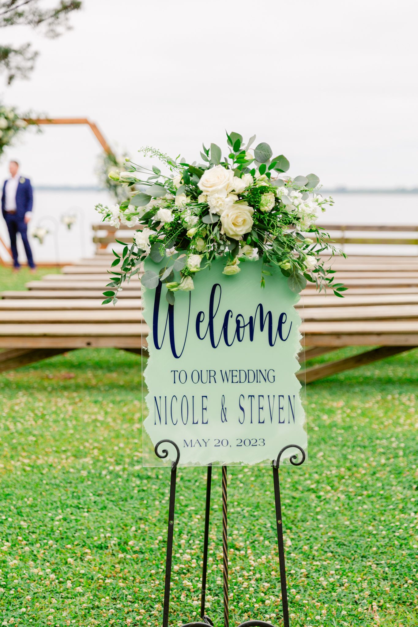 Elegant wedding welcome sign with white floral arrangement and greenery displayed on metal stand by waterfront.