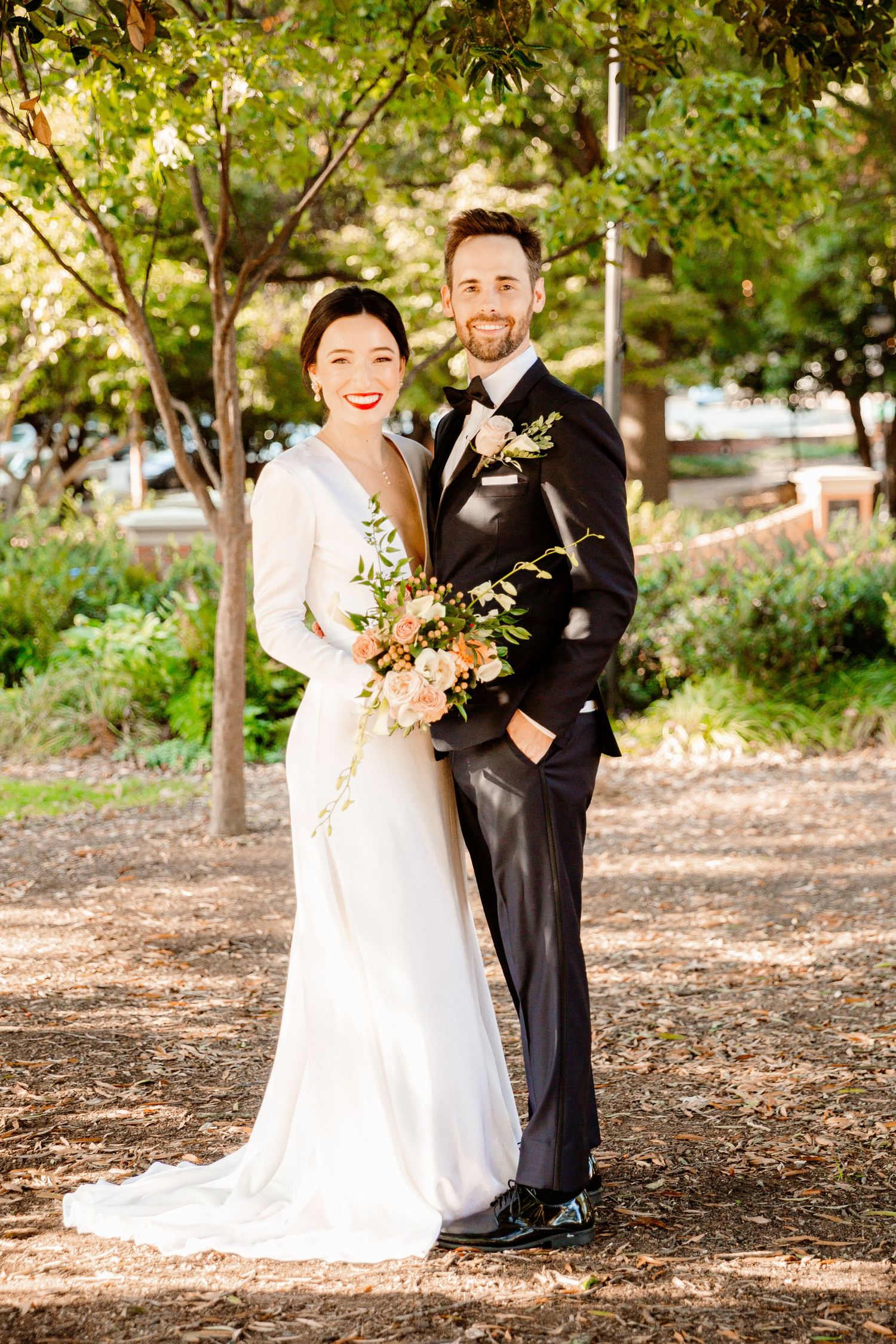A couple in wedding attire poses together in a sunlit outdoor garden setting with trees in the background.