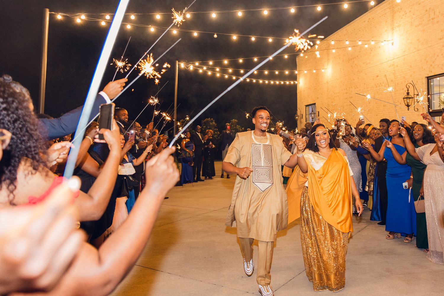 Guests hold sparklers creating a festive exit pathway at an evening wedding celebration under market lights.