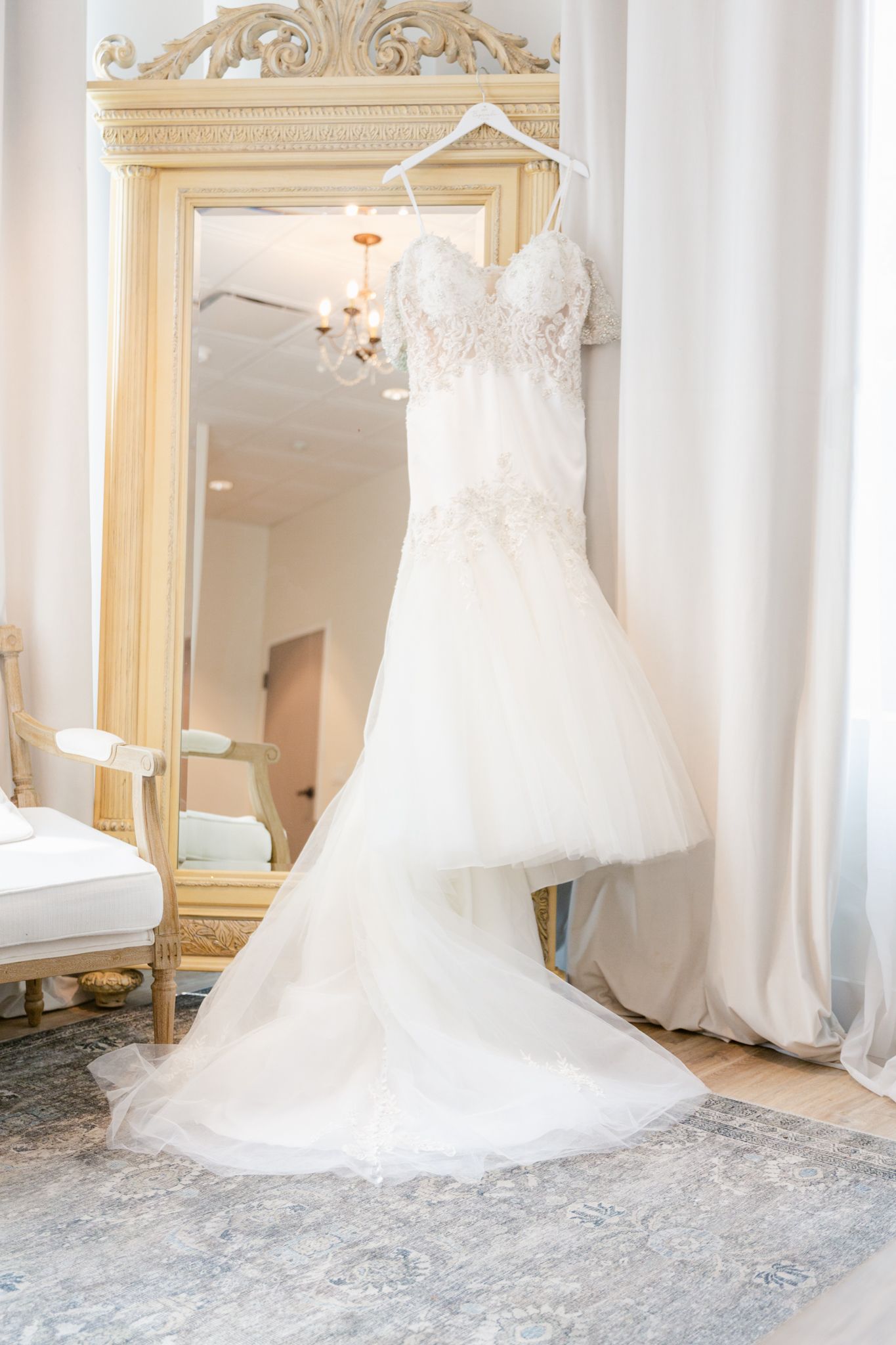 White lace wedding dress hanging on ornate gold mirror frame in a bridal suite.