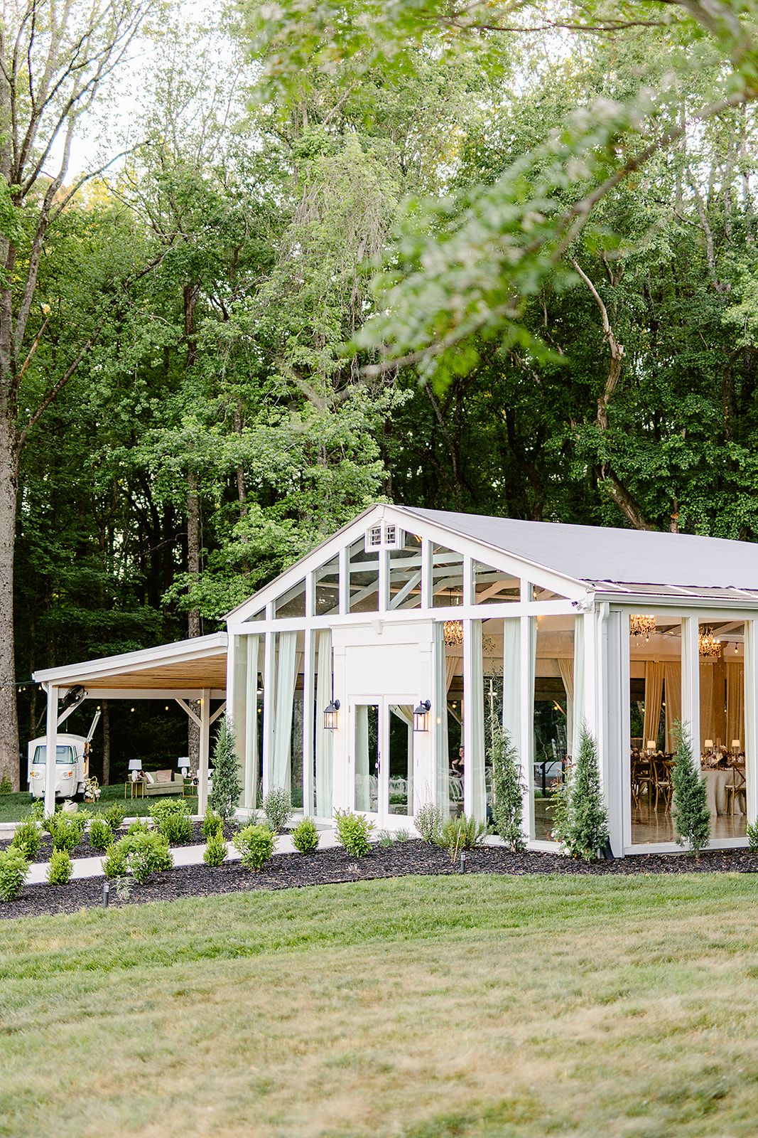 Modern greenhouse-style wedding venue with white frame and glass panels nestled in a wooded setting.