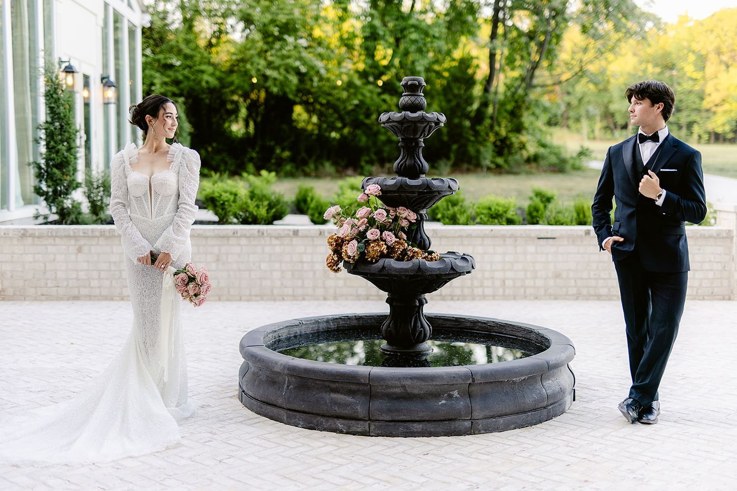 A bride and groom walk hand in hand through a garden path with a decorative fountain and lush greenery.