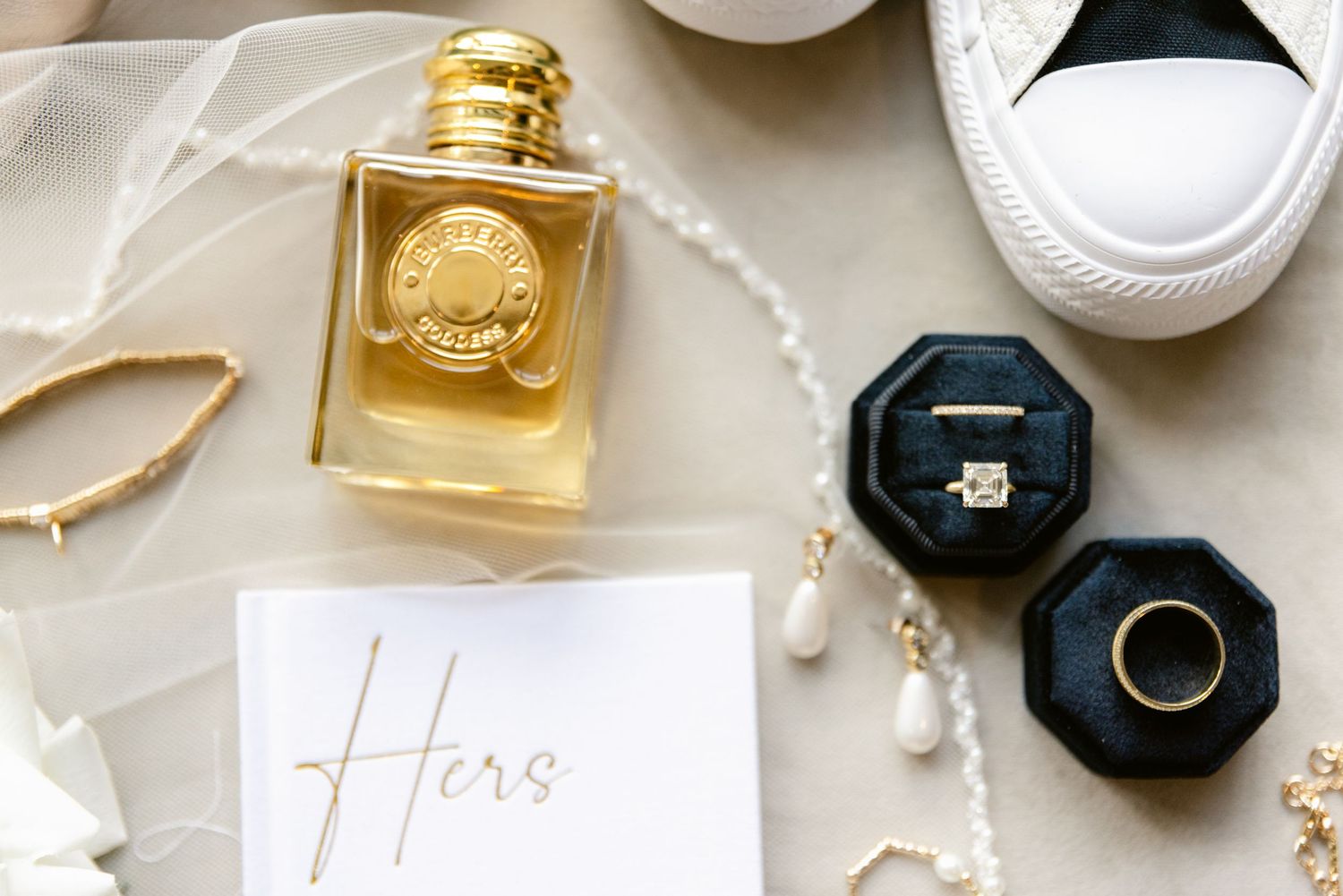 Wedding details flatlay with gold perfume bottle, pearl necklace, engagement ring box and white sneaker.