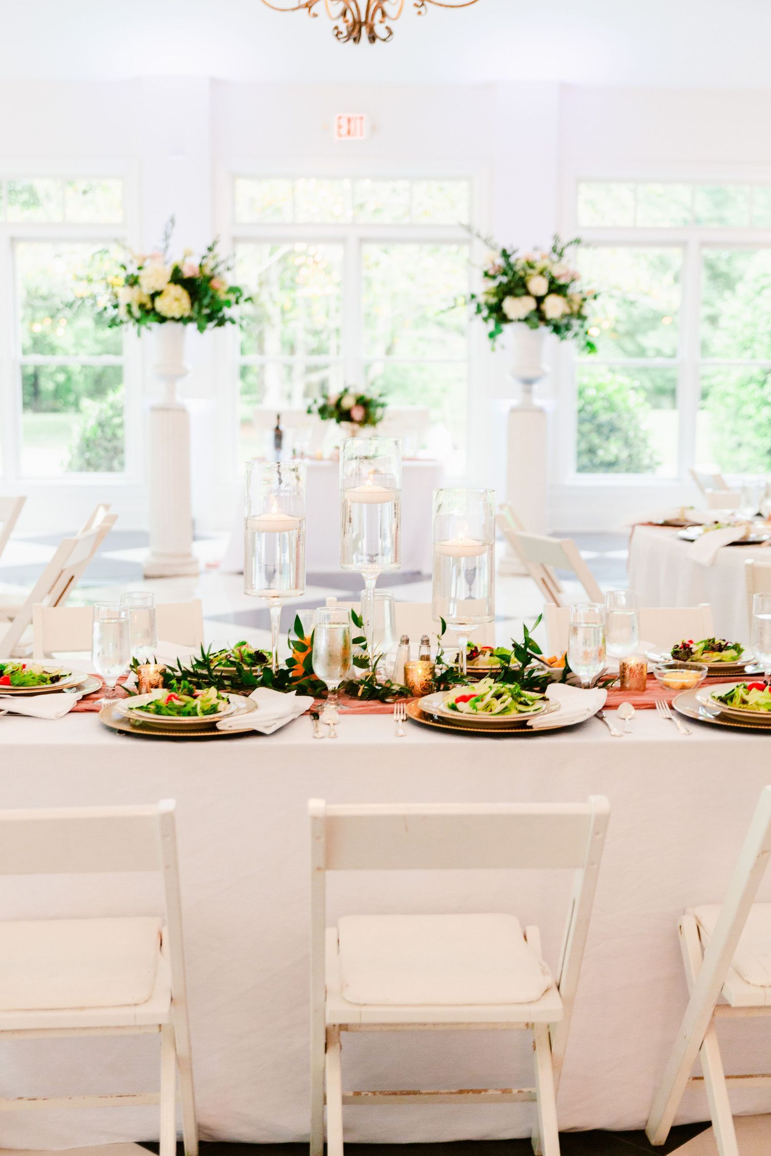 Elegant white wedding reception tables decorated with greenery garlands and gold charger plates in a bright venue.