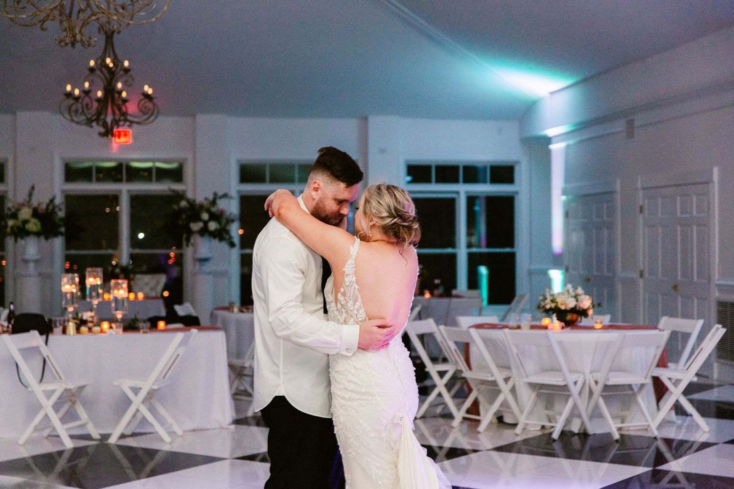A couple shares their first dance at an elegant wedding reception with white chairs and soft blue lighting.