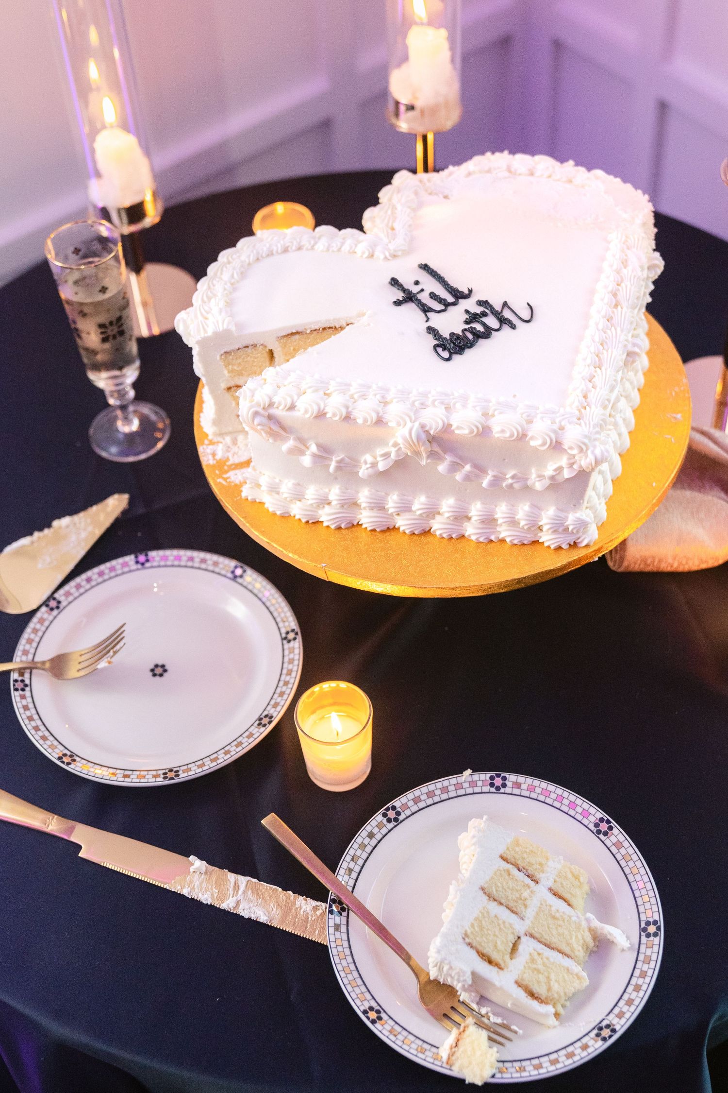 A heart-shaped white wedding cake with delicate piping sits among elegant table settings and lit candles.