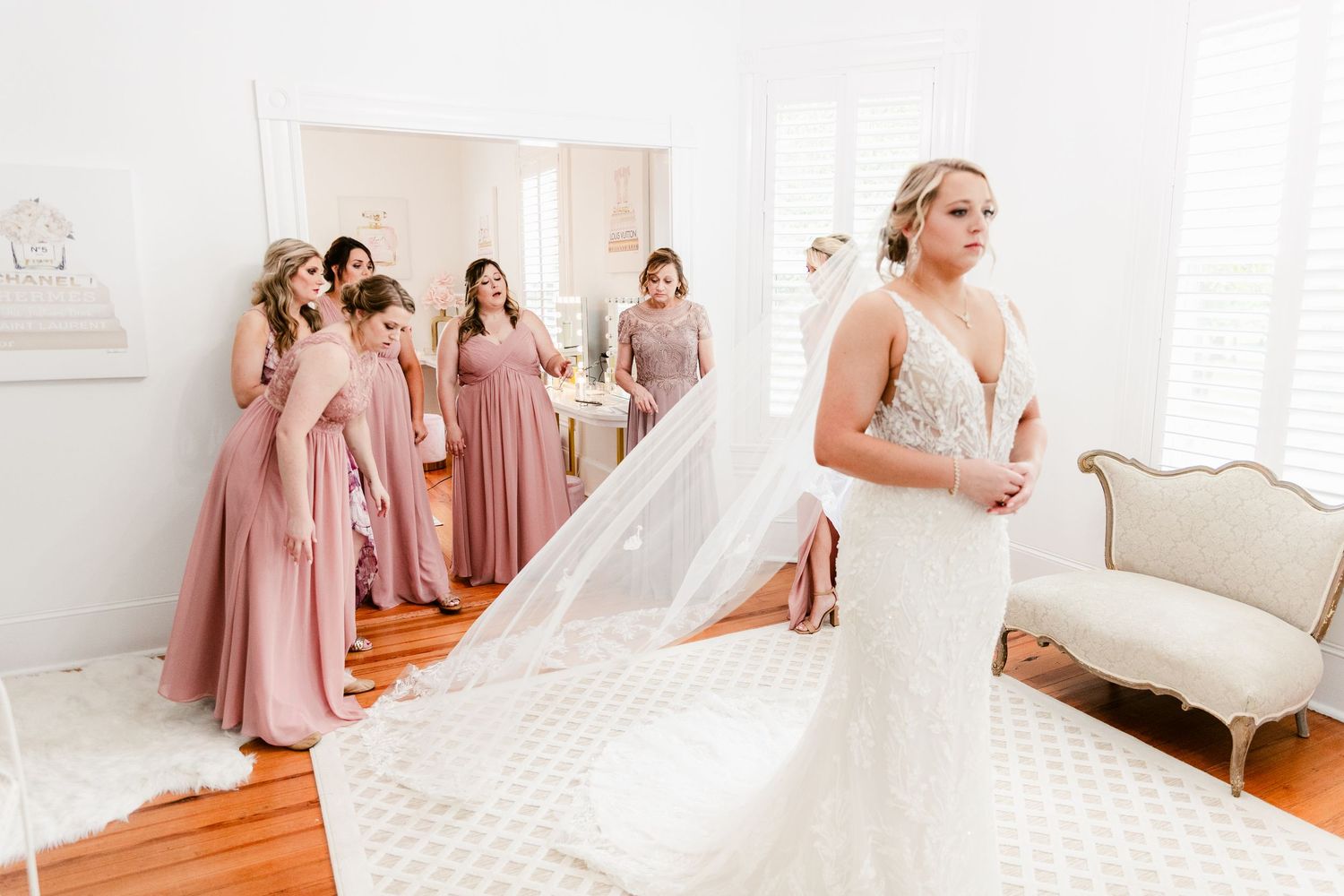 Bride and bridesmaids in dusty rose dresses share candid moments while getting ready in a bright white room.