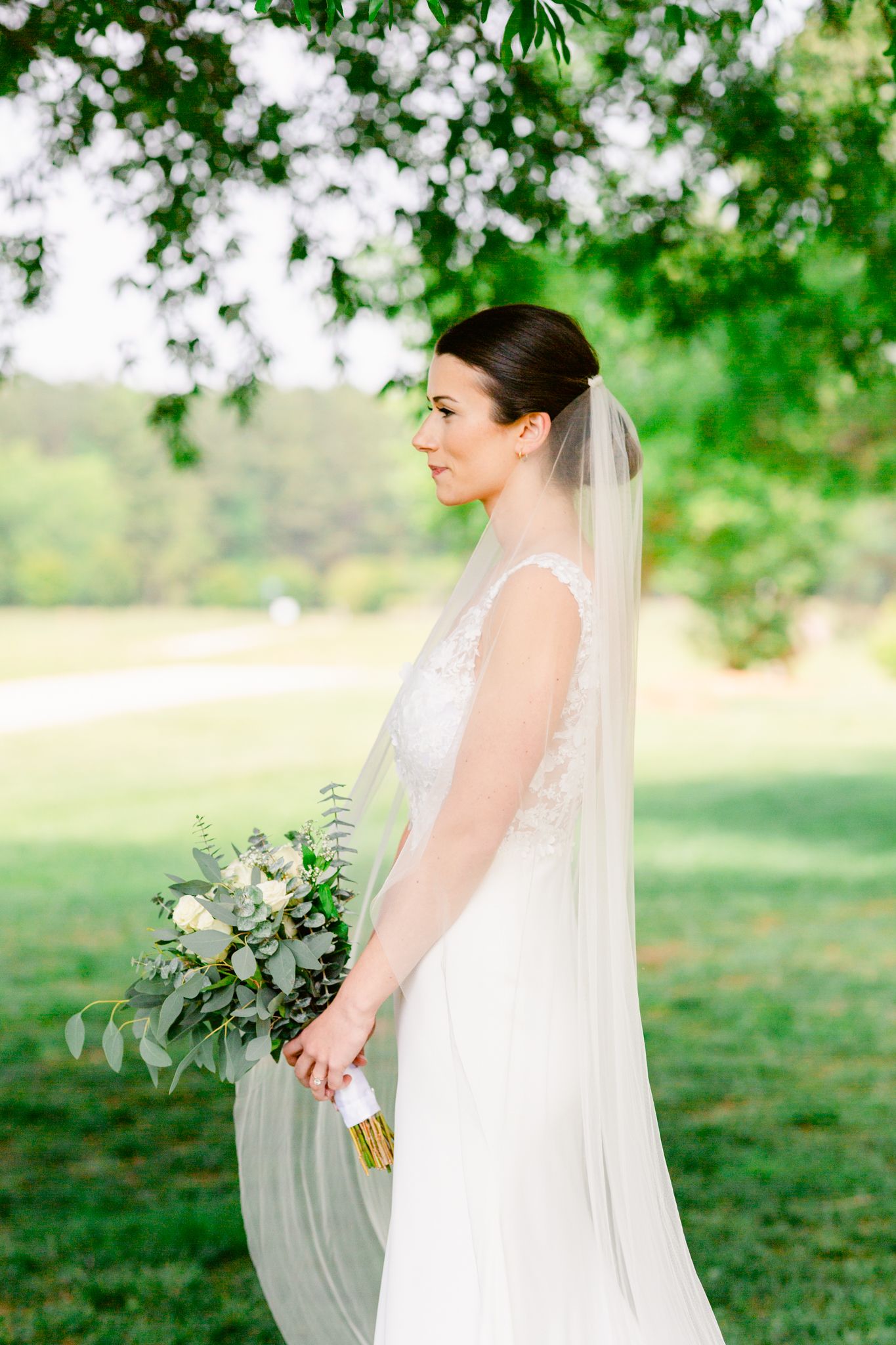 A bride in a flowing white gown and veil holds a bouquet while standing in a sunlit garden setting.