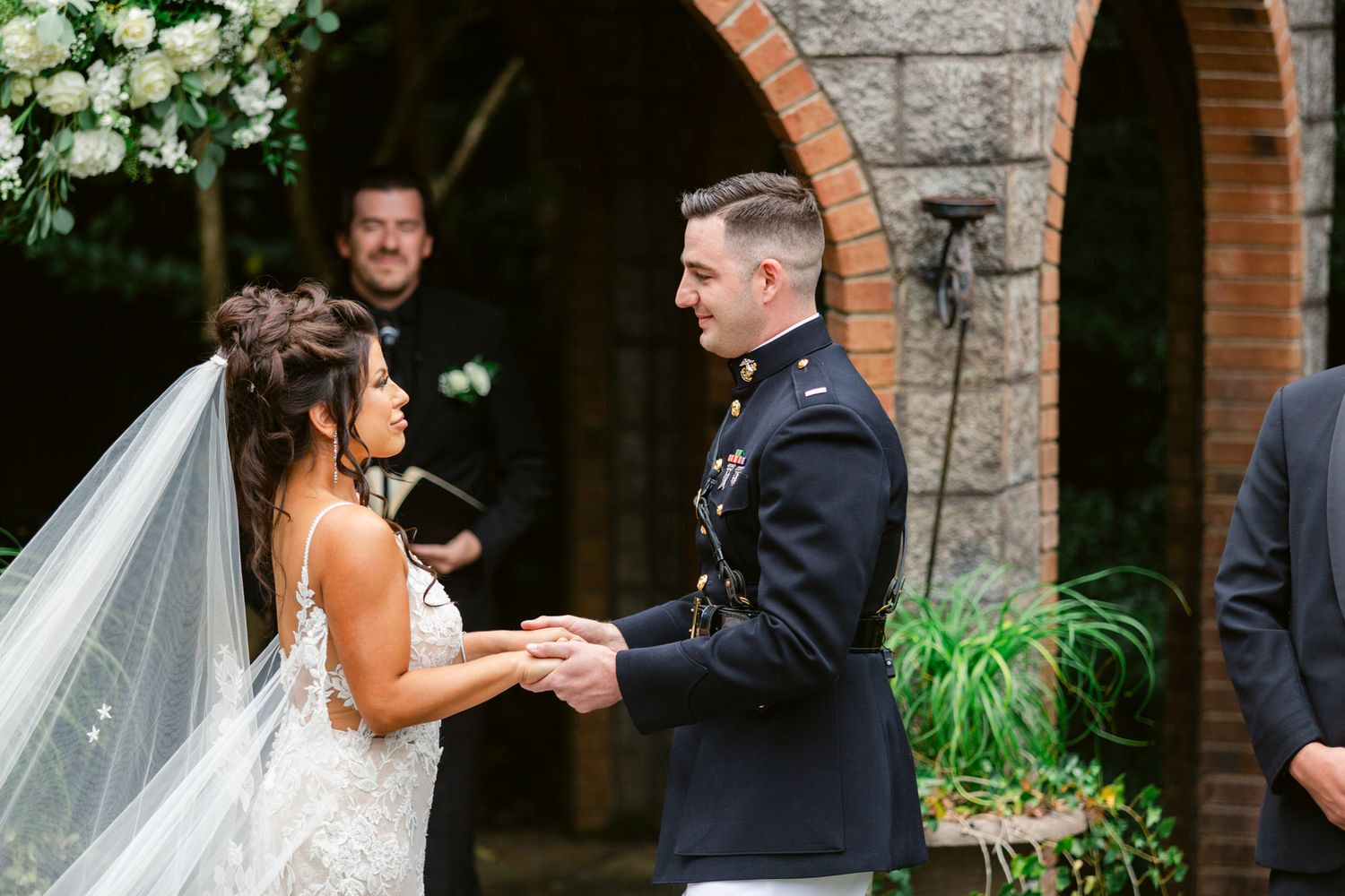 A romantic wedding ceremony takes place in a rustic stone courtyard with greenery and white floral decorations.