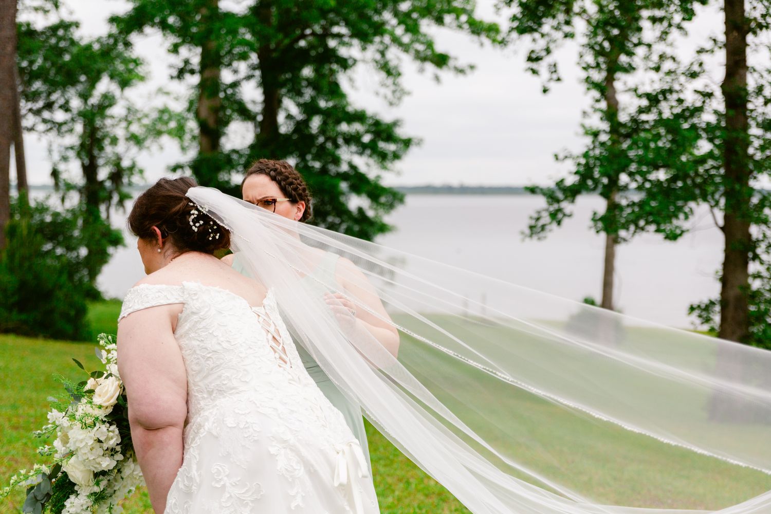 A couple in wedding attire embraces on a lush green lawn overlooking a serene lake on their special day.