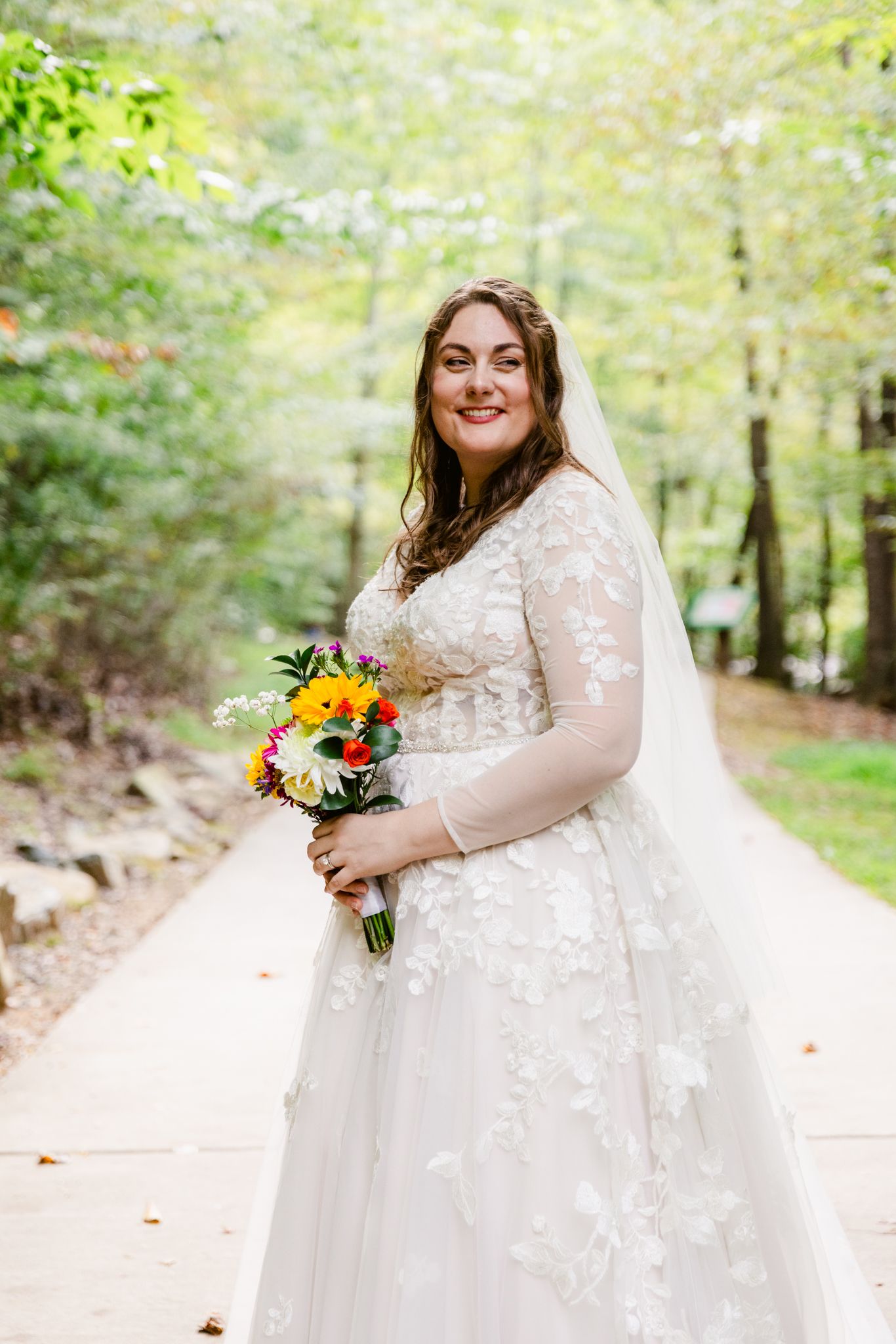 A bride in a lace sleeve wedding gown holds a vibrant sunflower bouquet while posing outdoors in a wooded setting.