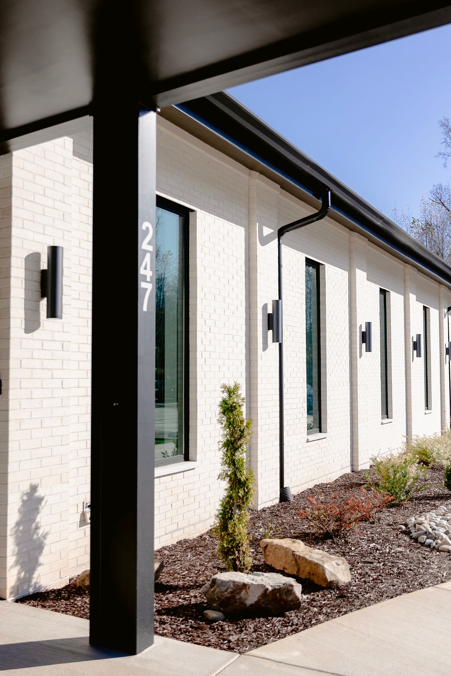 White exterior building facade with black columns and landscaped mulch beds along a covered walkway.
