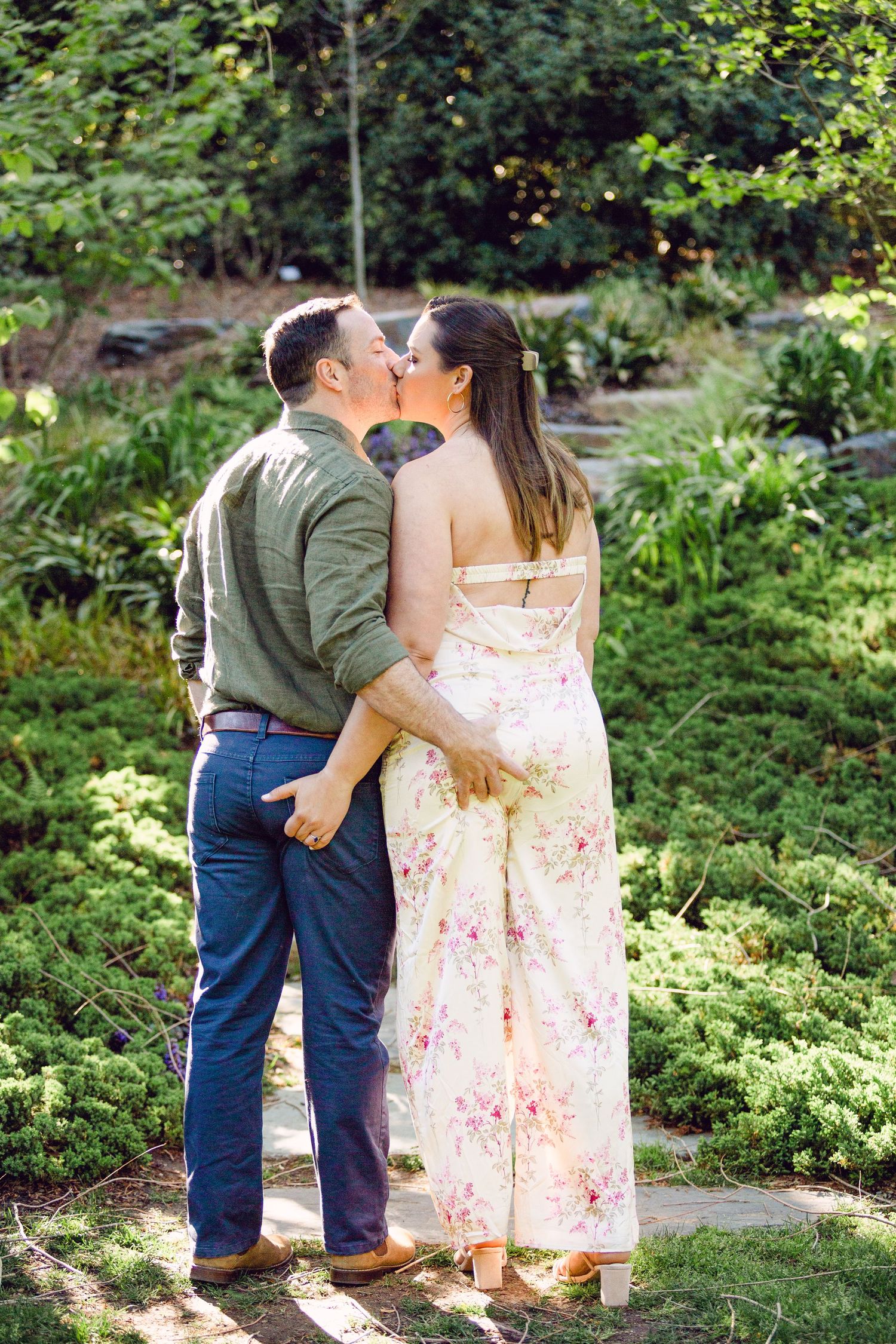 A romantic couple shares tender moments in a lush garden setting with ferns and greenery surrounding them.