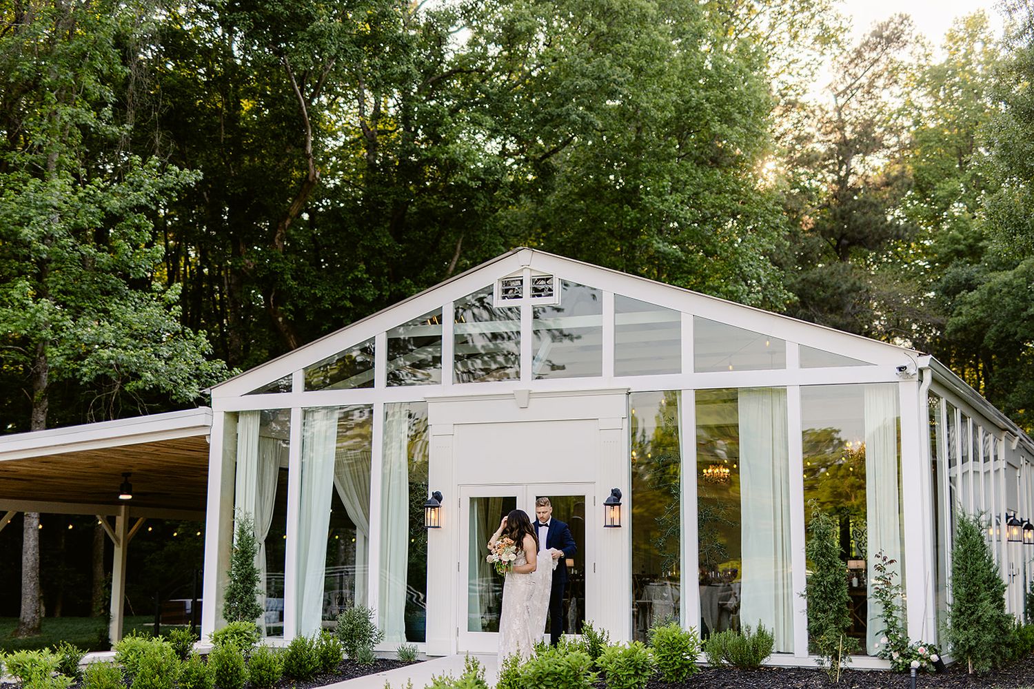 Row of white outdoor garden pavilions with glass windows nestled among lush green trees in a tranquil setting.