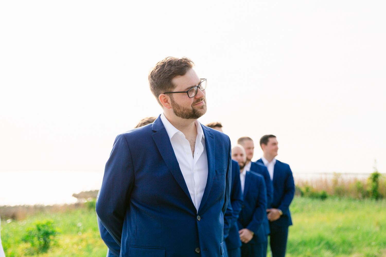 Wedding party in navy blue suits walks through a green field on a sunny summer day.