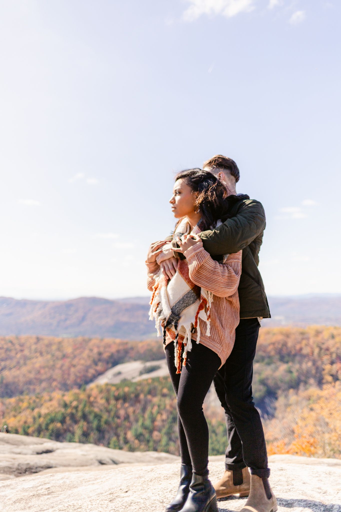 A couple embraces while standing on a mountain overlook with autumn foliage and rolling hills in the background.