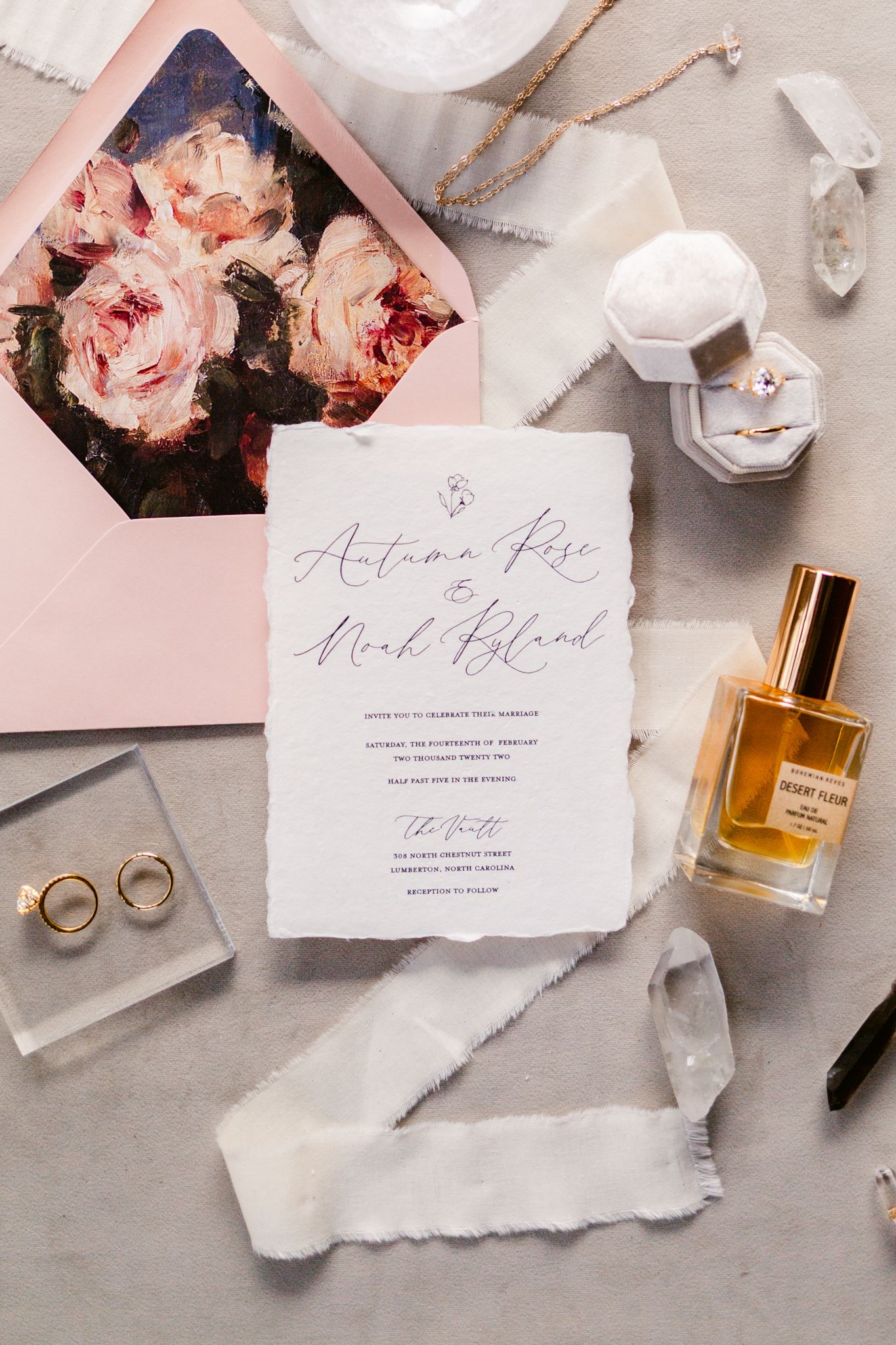 Elegant wedding invitation flatlay with torn paper edges, pink roses, perfume bottle and gold rings on gray surface.