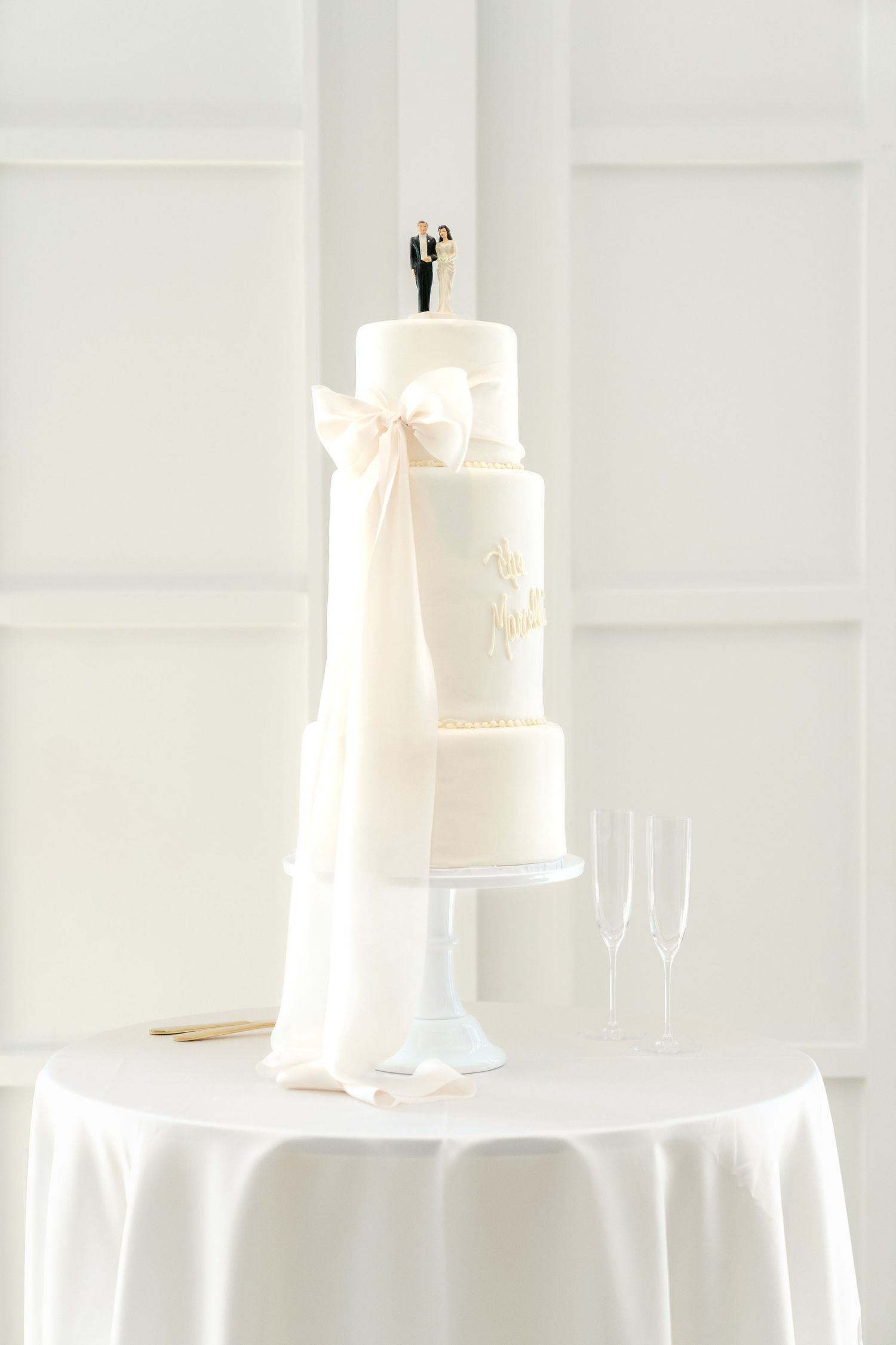 Elegant white wedding cake with multiple tiers displayed on white tablecloth with champagne glasses in background.