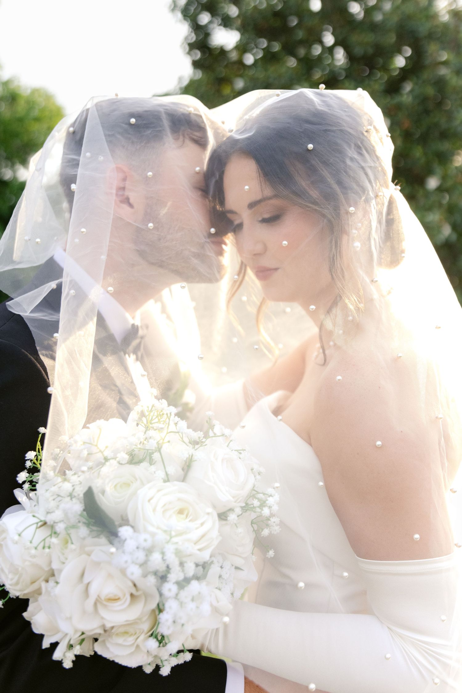 A romantic wedding portrait under sunlight with a veil and elegant white rose bouquet.