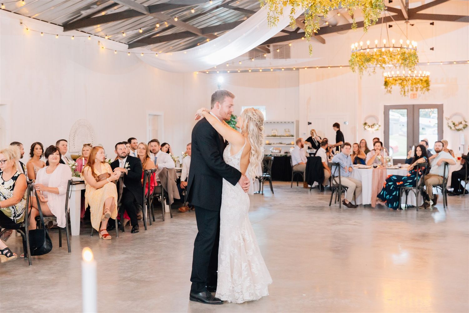 Wedding couple dances in modern reception space with guests watching from tables.