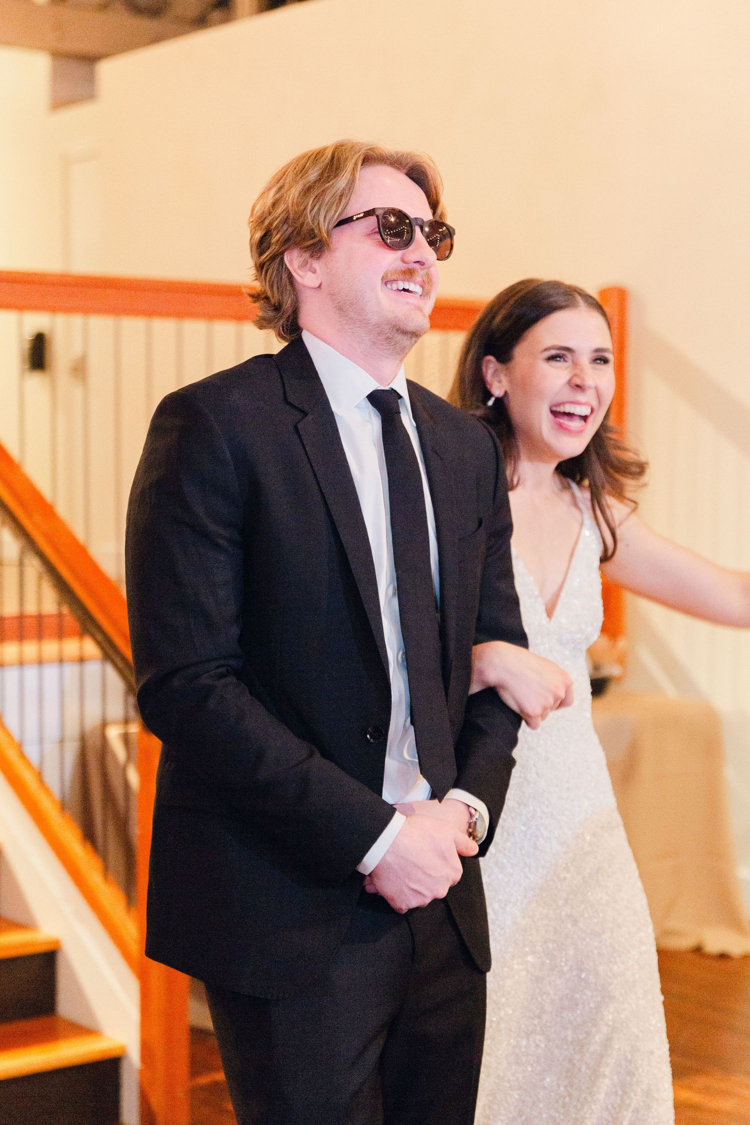 A couple sharing a candid laughing moment while standing in a bright indoor venue with wooden stairs.