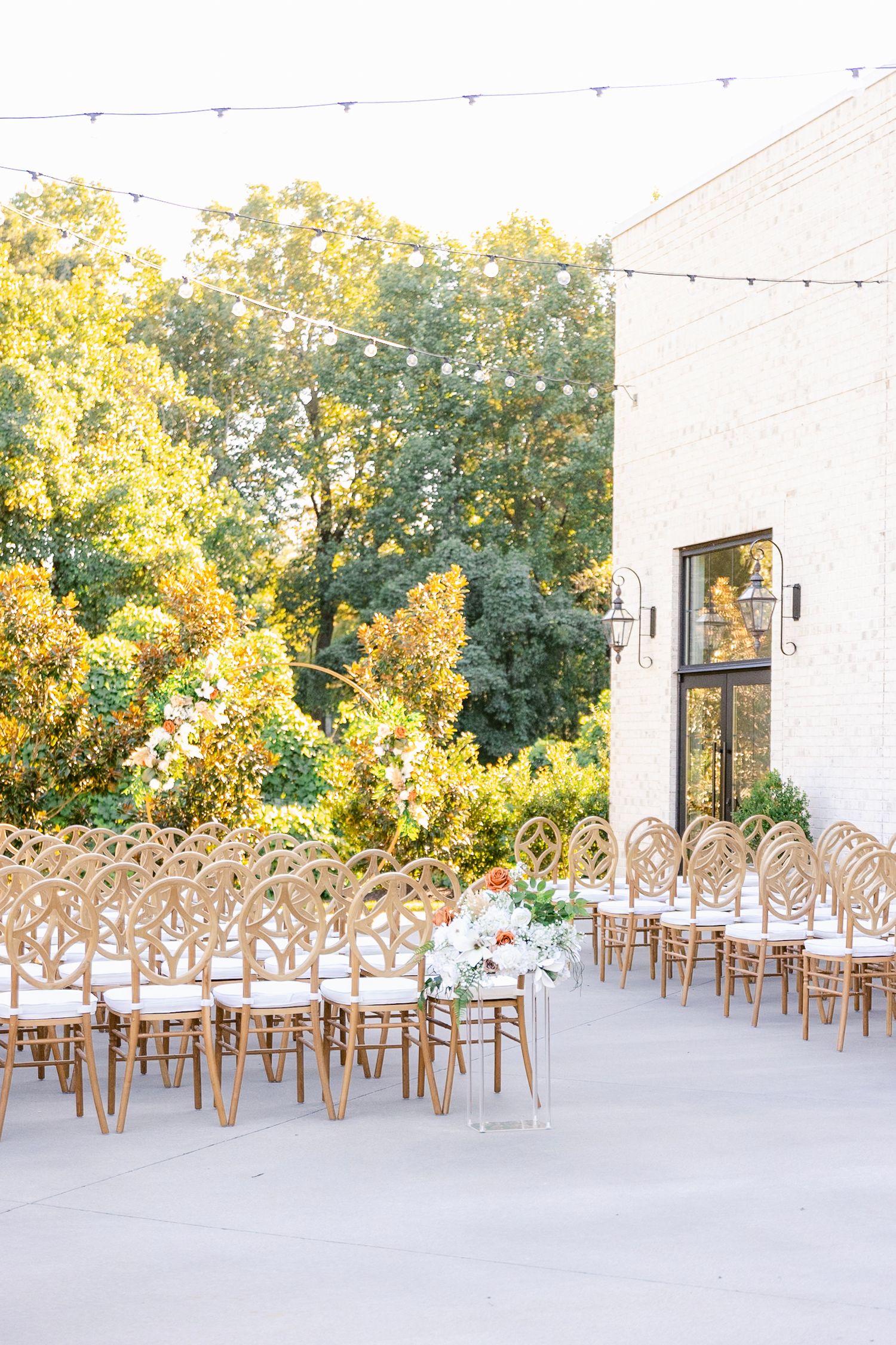 Outdoor wedding ceremony setup with wooden chairs arranged on a patio surrounded by autumn trees.