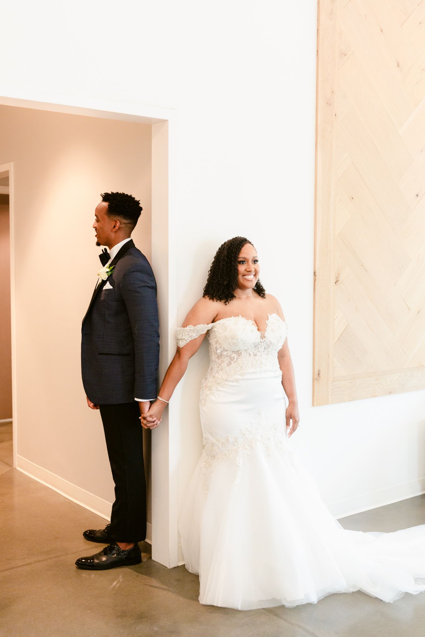 A couple in wedding attire shares a playful first look moment around a corner in a bright indoor venue.