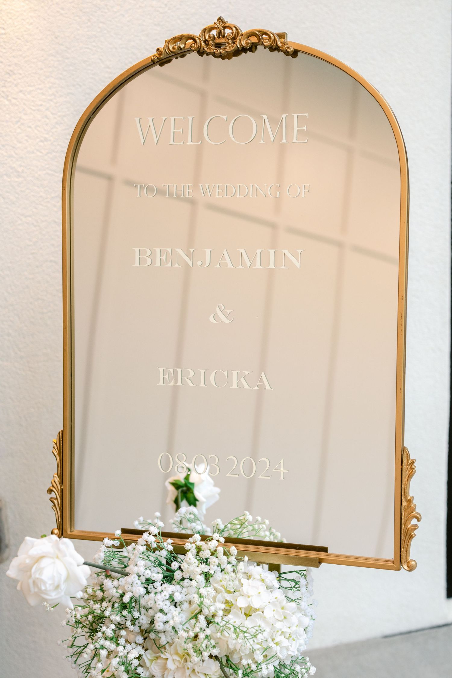 Elegant gold-framed mirror with white floral decorations for a wedding welcome display.