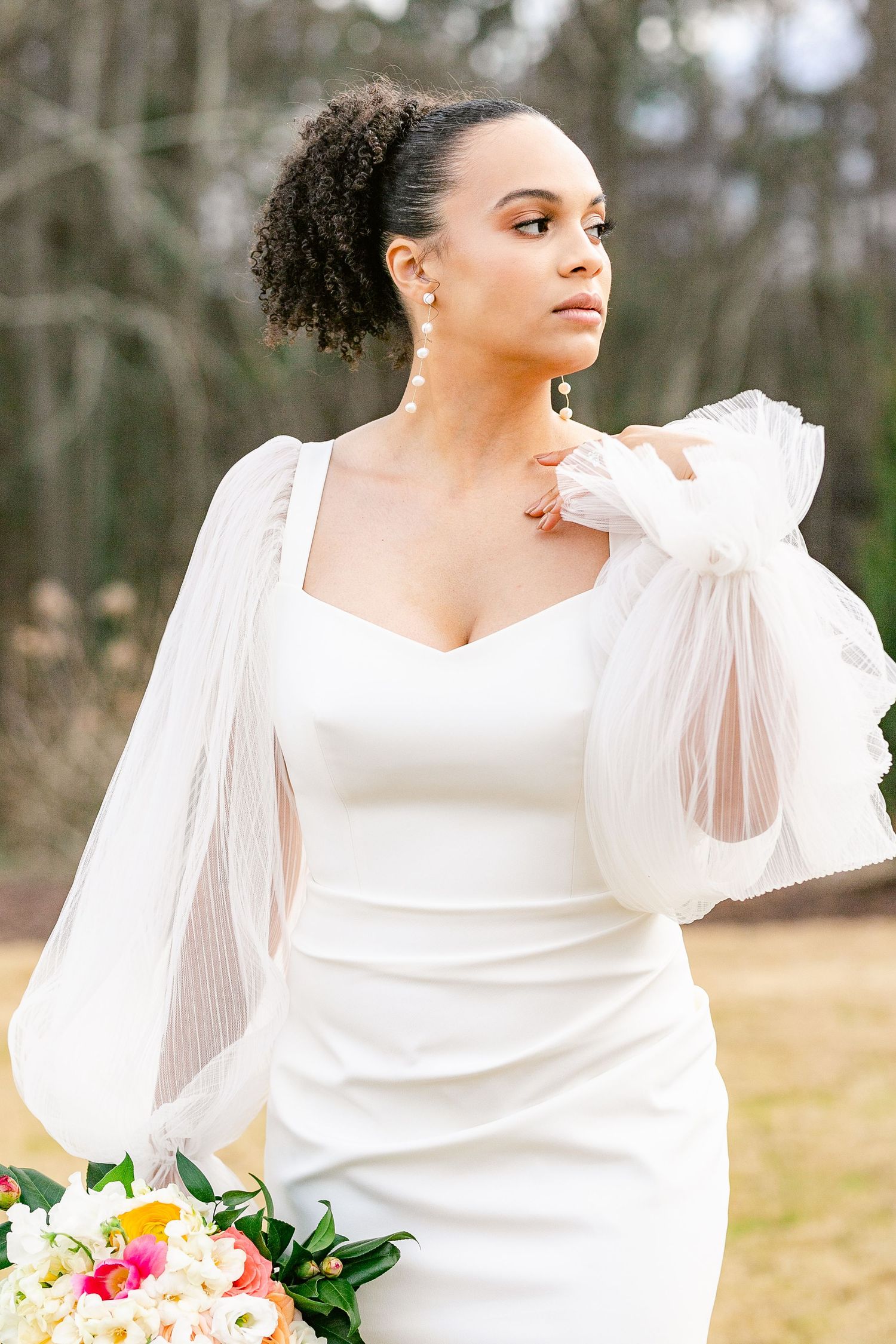 A bride in a modern white dress with dramatic puff sleeves poses elegantly against a natural outdoor backdrop.