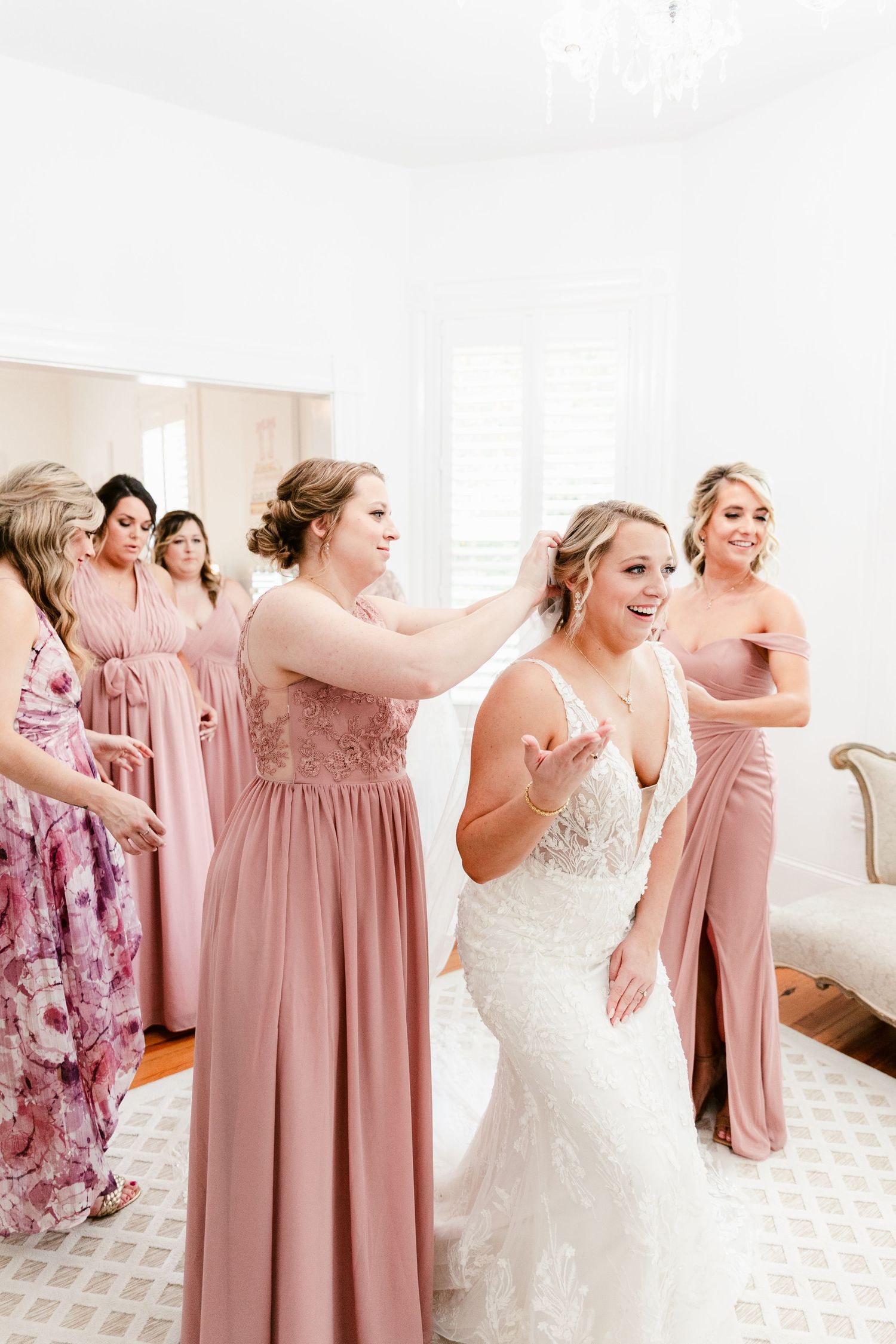 A bridal party in dusty rose bridesmaid dresses and a white wedding gown laughs together in a bright room.