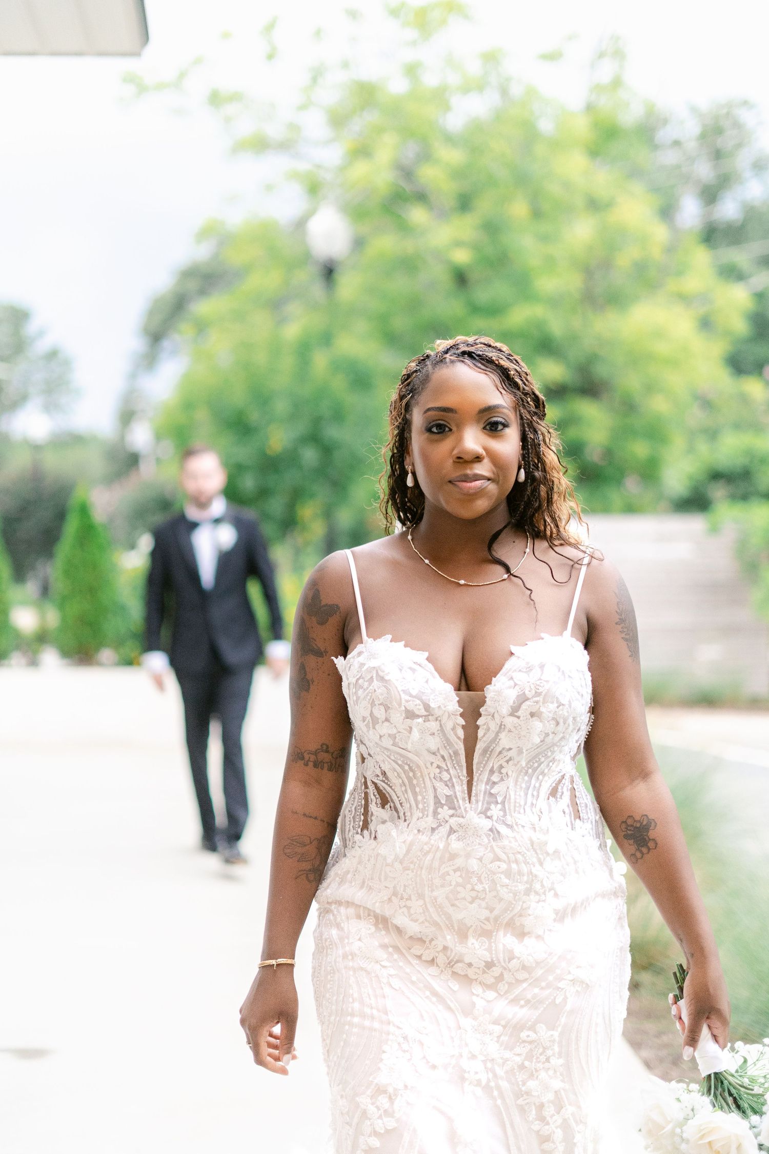 A bride in a white lace wedding dress poses outdoors in a garden setting with trees in the background.